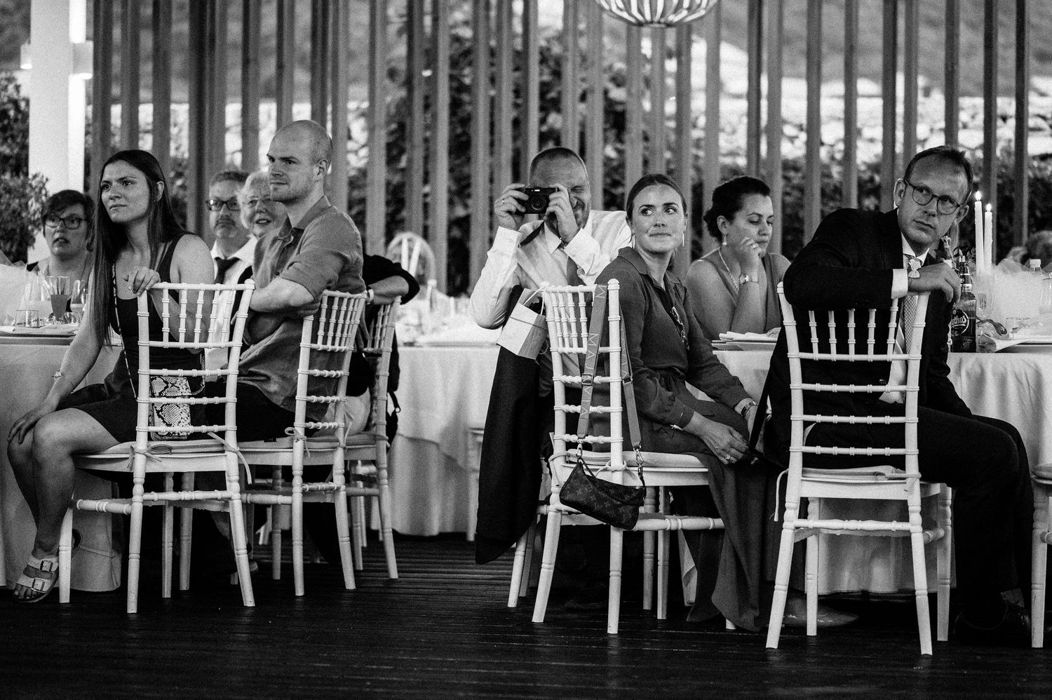 Wedding guests seated at long tables with white chairs during an outdoor reception.