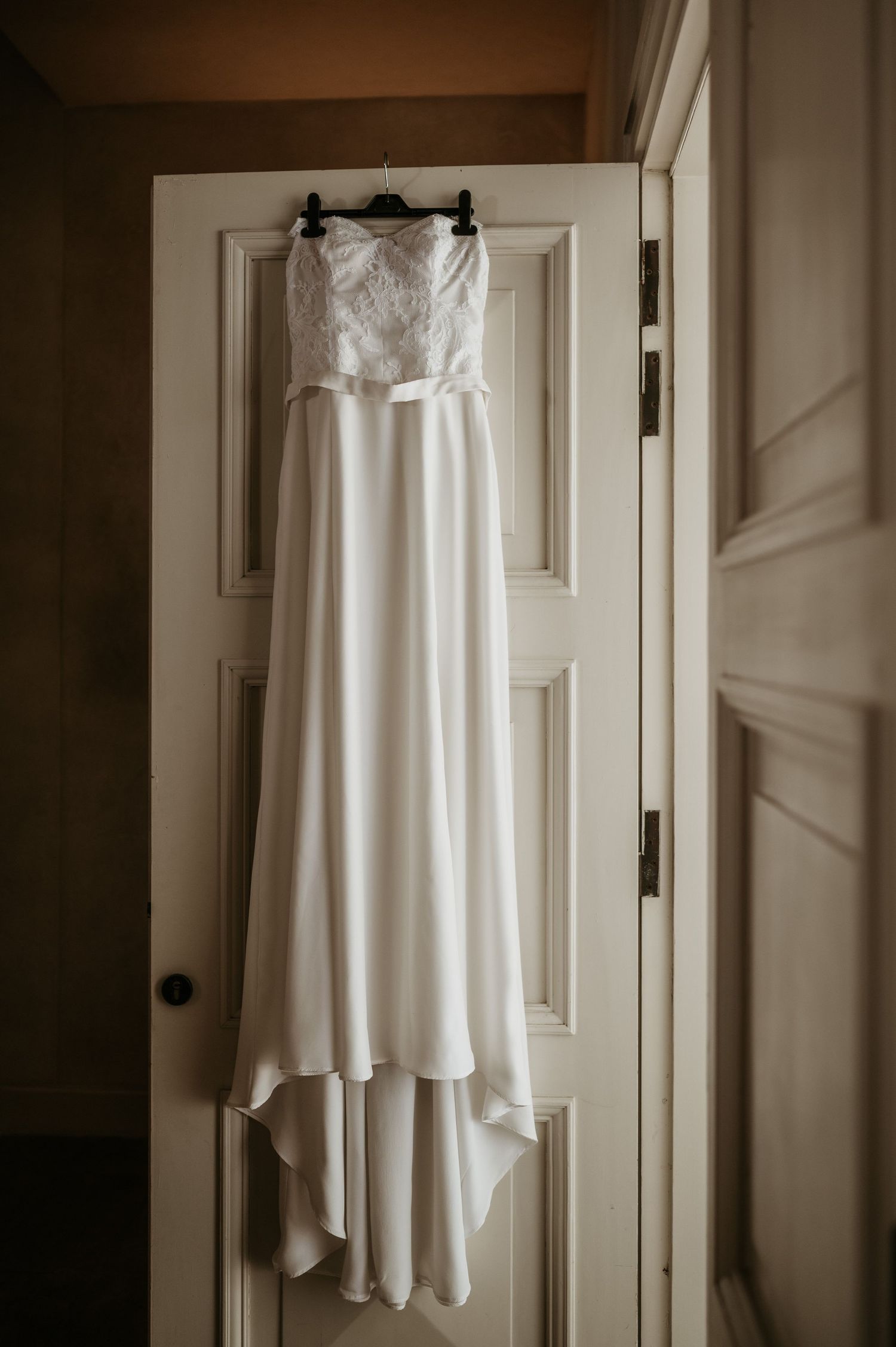 An elegant white wedding dress with lace detailing hangs on a door against a neutral wall.