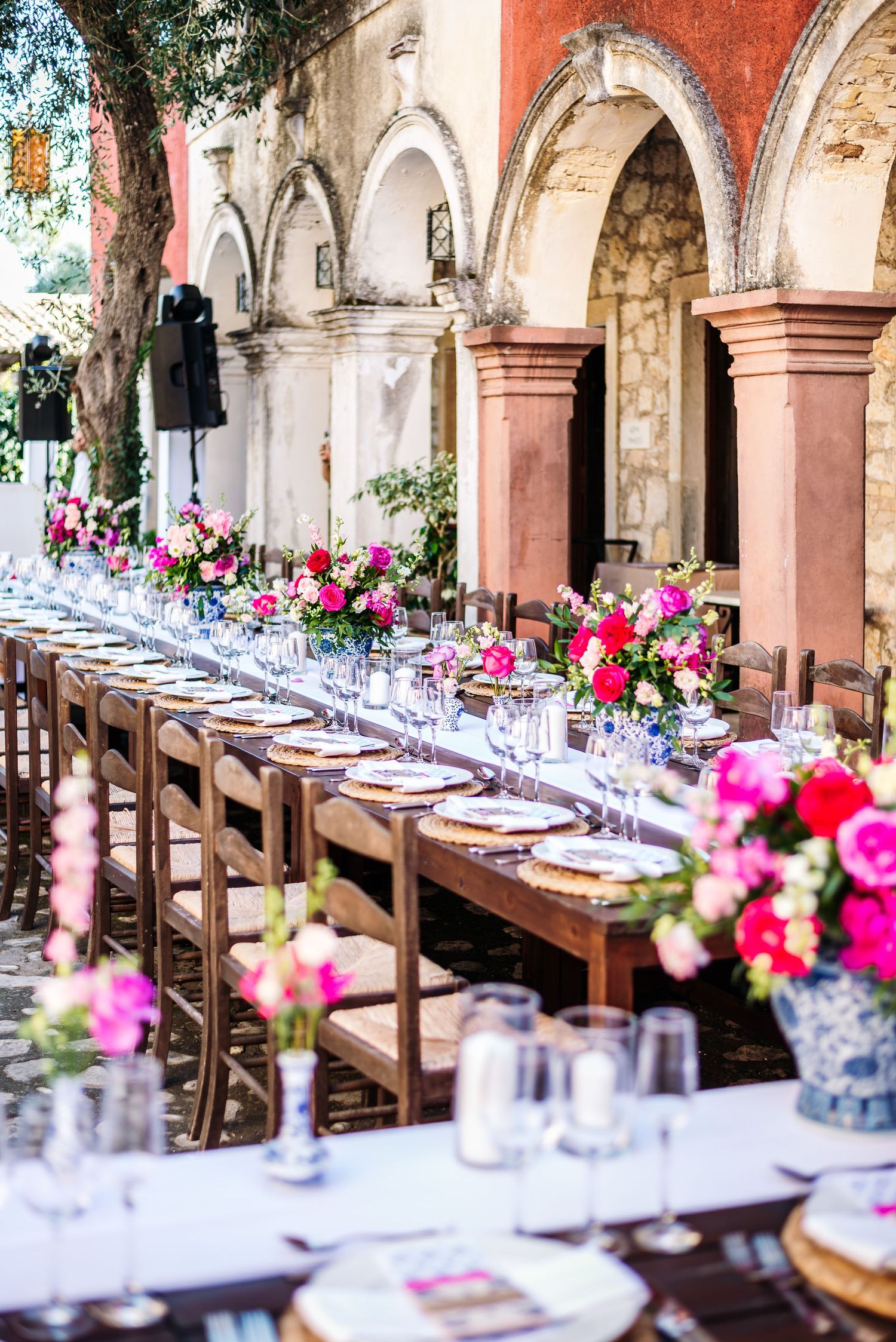 Long wedding reception table with pink floral centerpieces under arched Mediterranean colonnade.