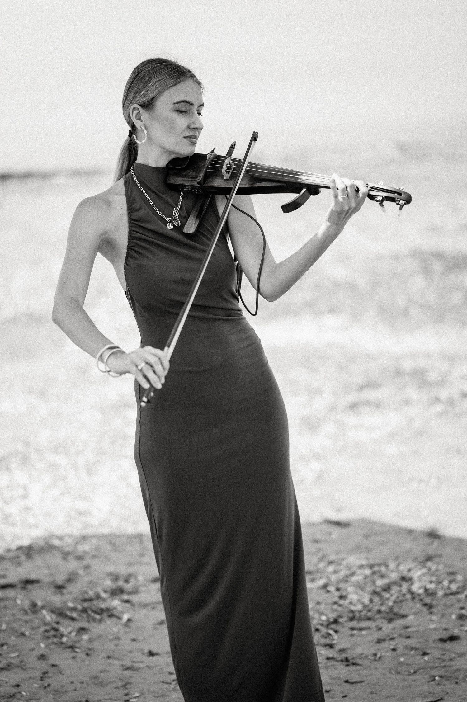 Violinist in a black dress performs on a beach in dramatic black and white photography.