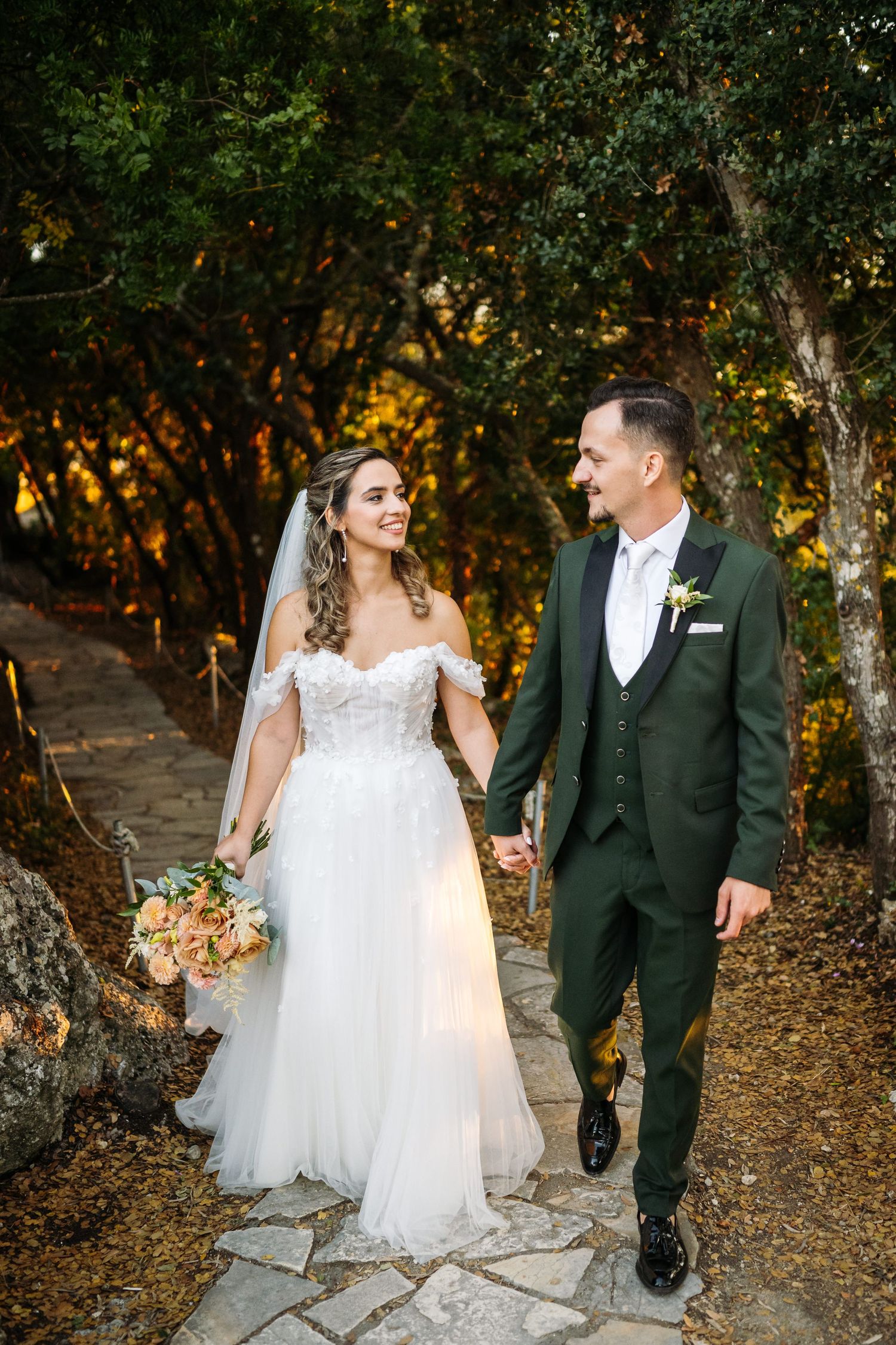 Newlyweds walking along stone path surrounded by lush greenery at sunset.