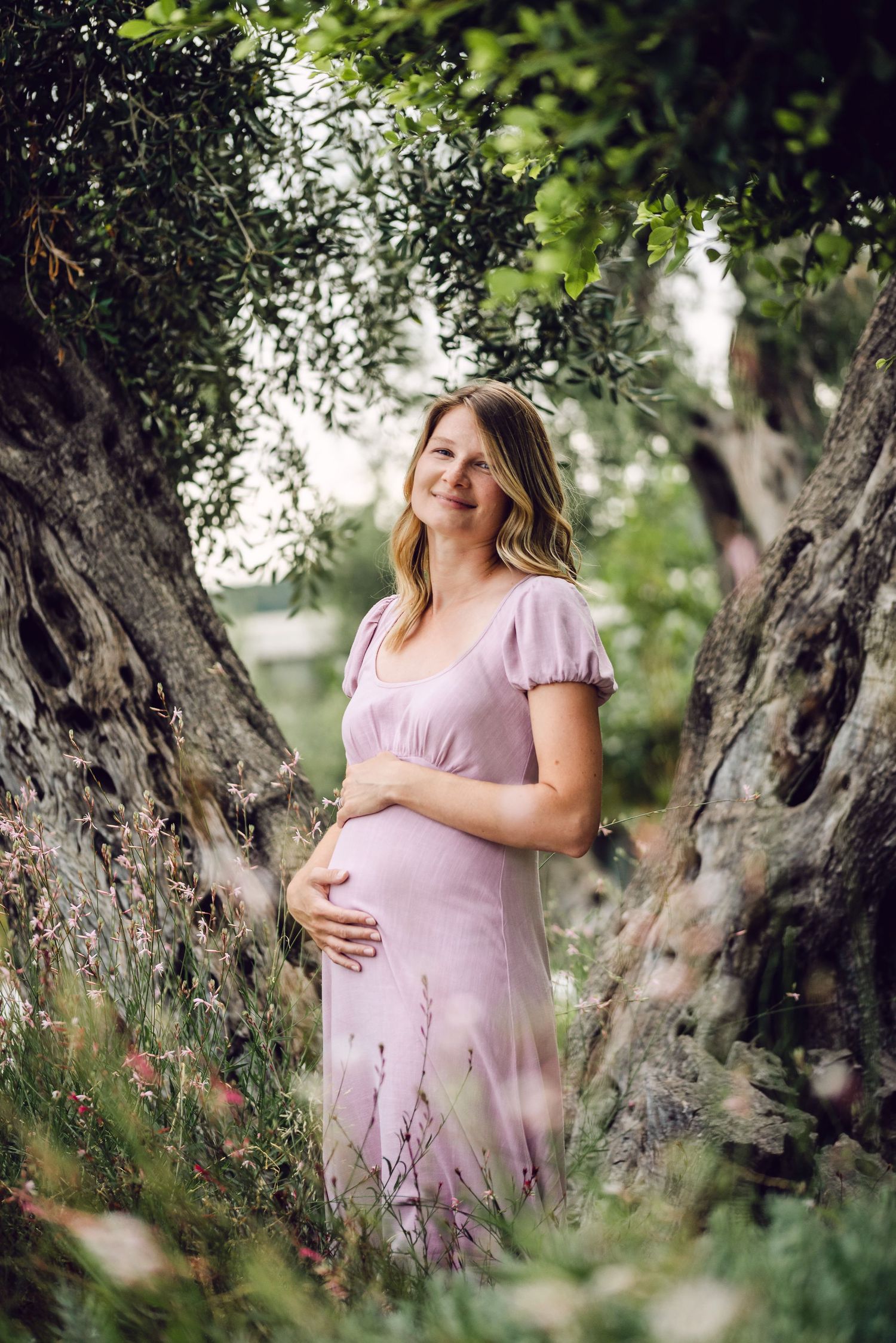 Maternity photos in a garden setting with olive trees showing a woman in a pink dress.