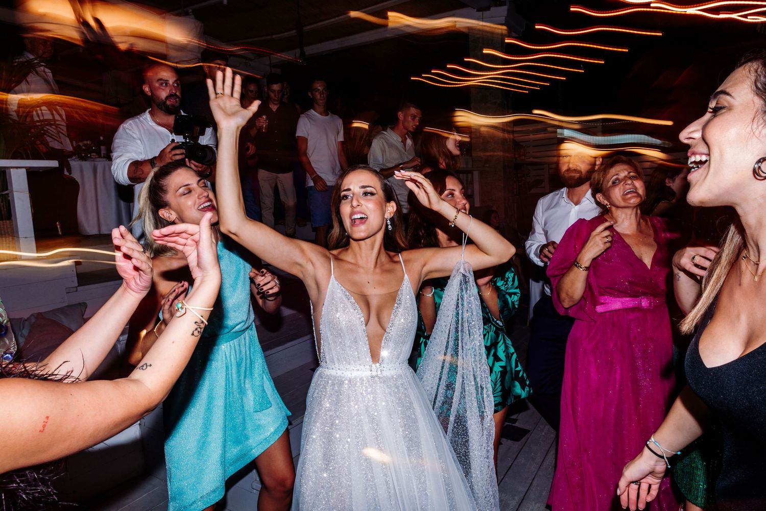 Wedding party dancing energetically during reception celebration with colorful lighting.