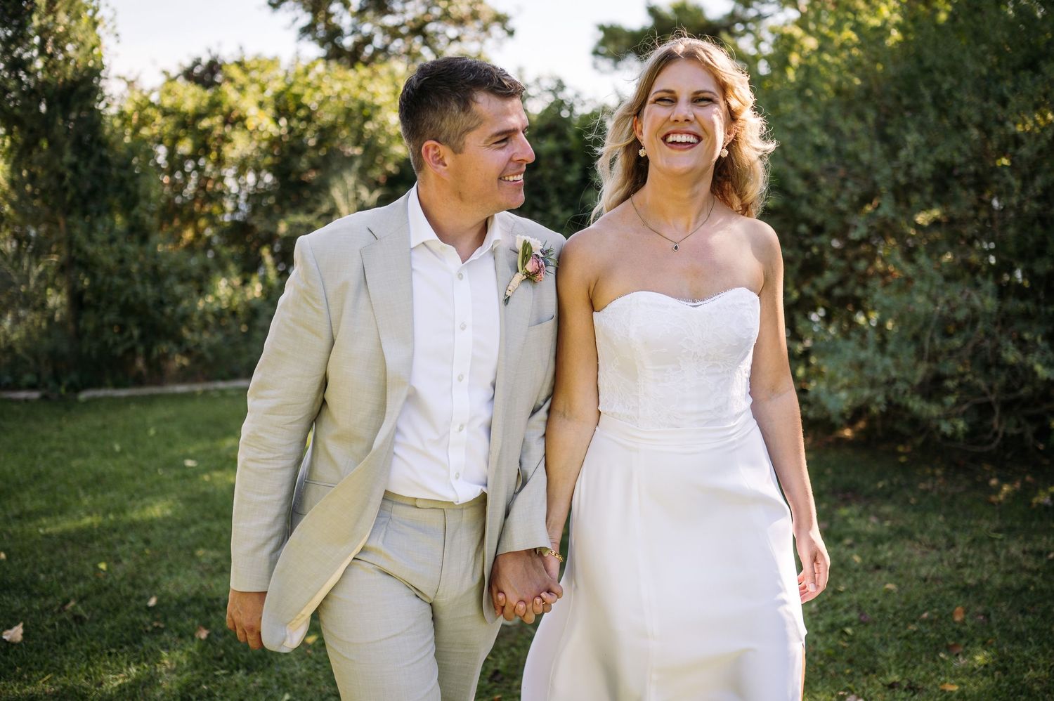 Couple in white and beige attire laugh while walking through garden setting.