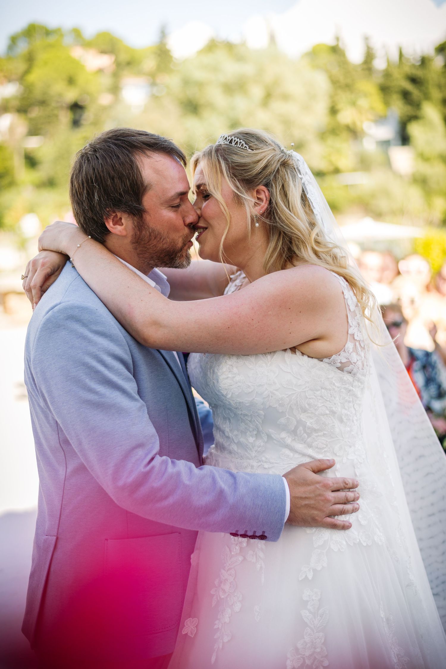 A tender wedding kiss captured during an outdoor ceremony with pink floral accents.