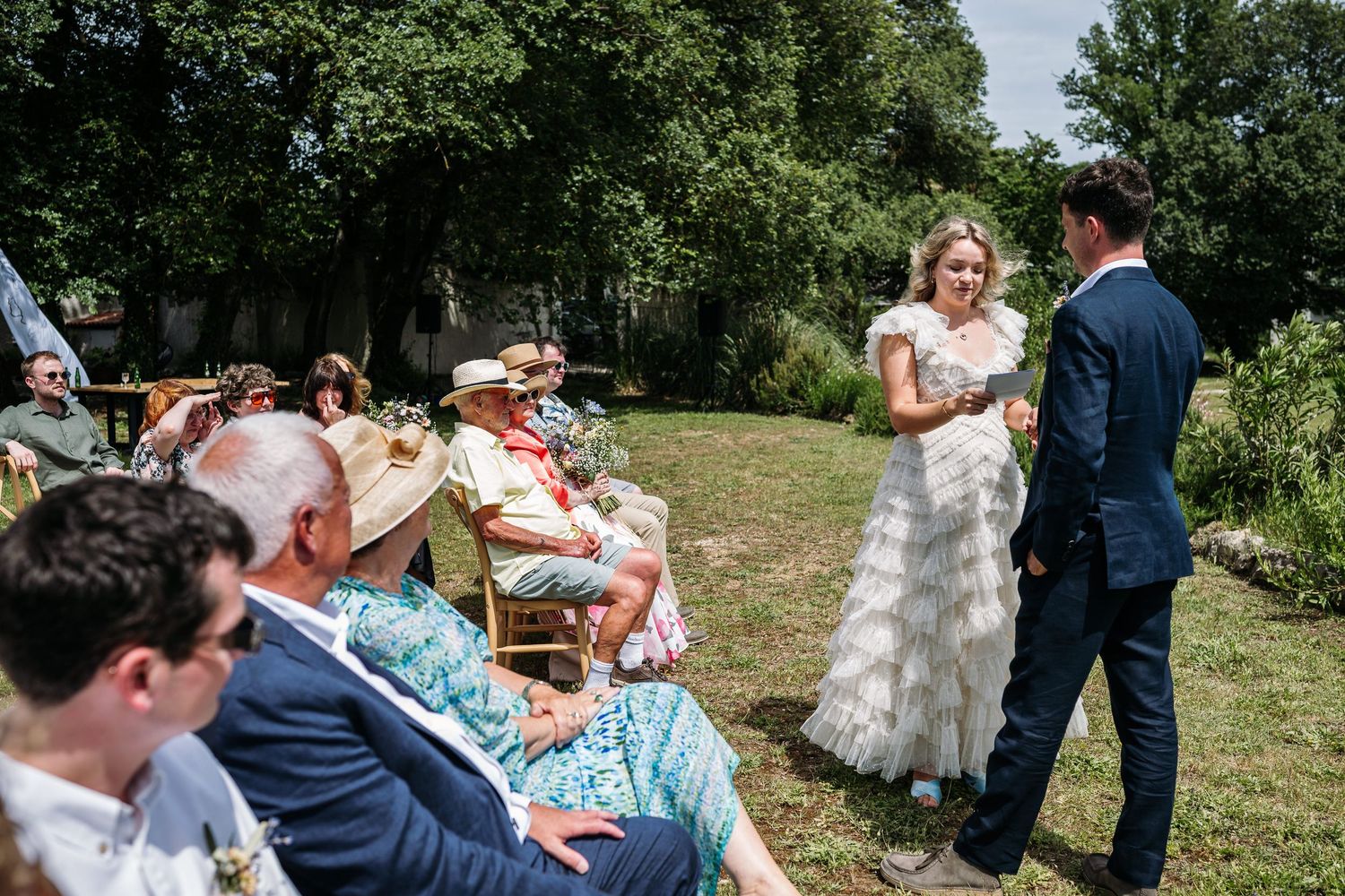 Wedding celebration in progress with guests seated on wooden chairs in a garden setting.