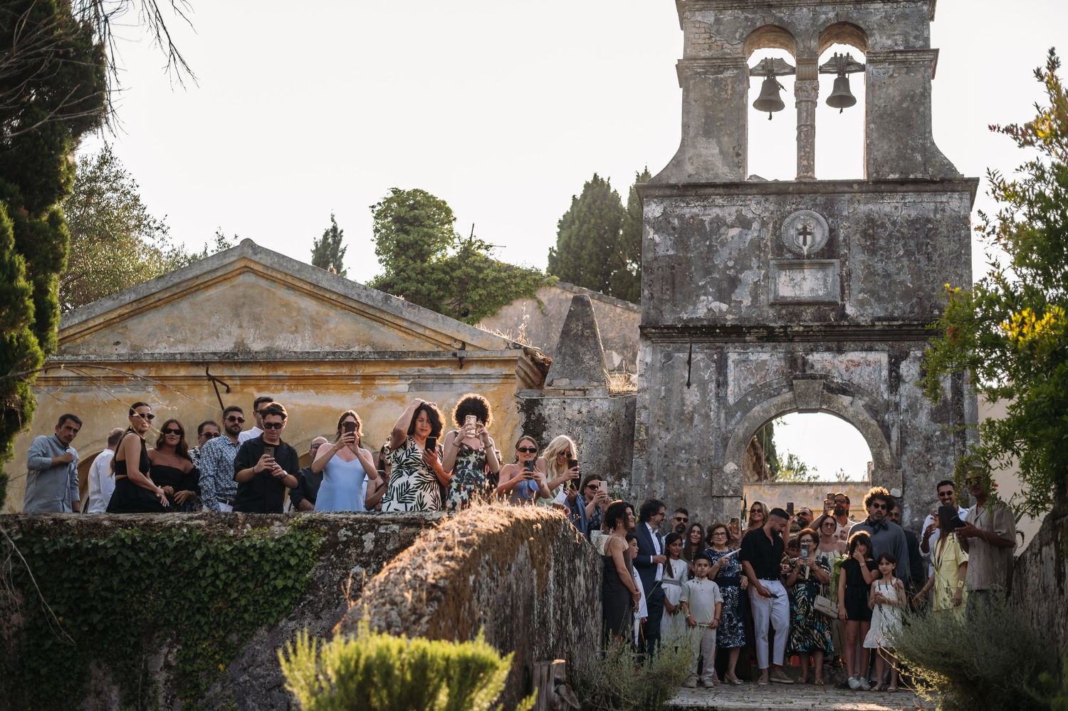 Large group of wedding guests gathered near historic stone bell tower at sunset.
