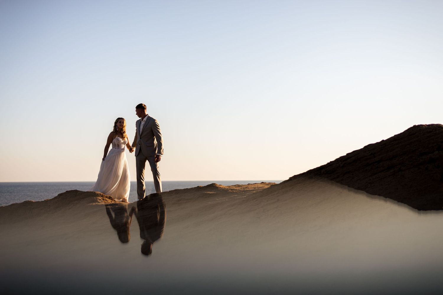 A dramatic sunset photo shows a wedding couple's reflection in calm coastal waters.