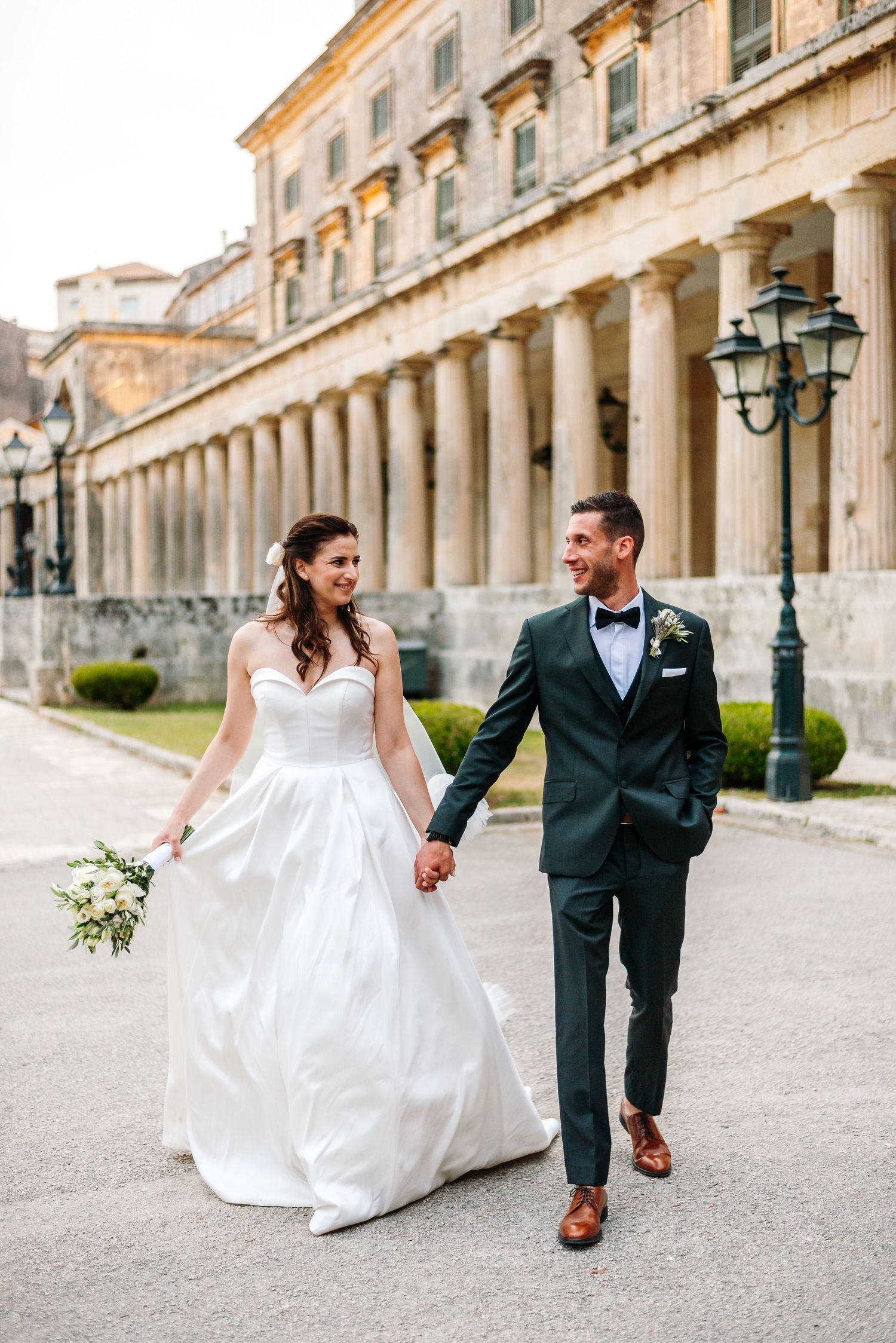 Newlyweds walk hand in hand along historic colonnade with beautiful architectural details.