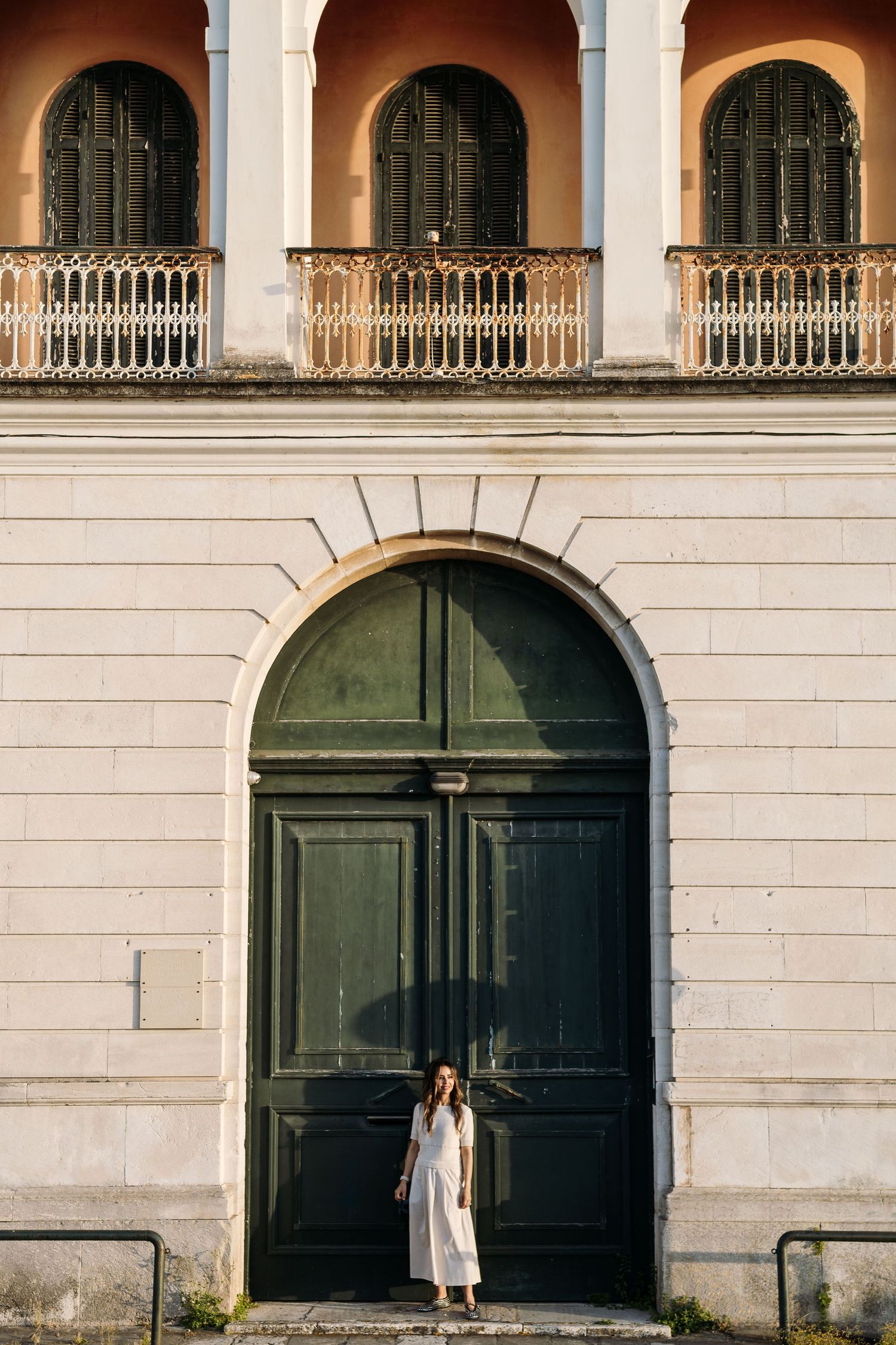 Person in white dress stands in front of large green doors beneath ornate arched balconies of historic building.