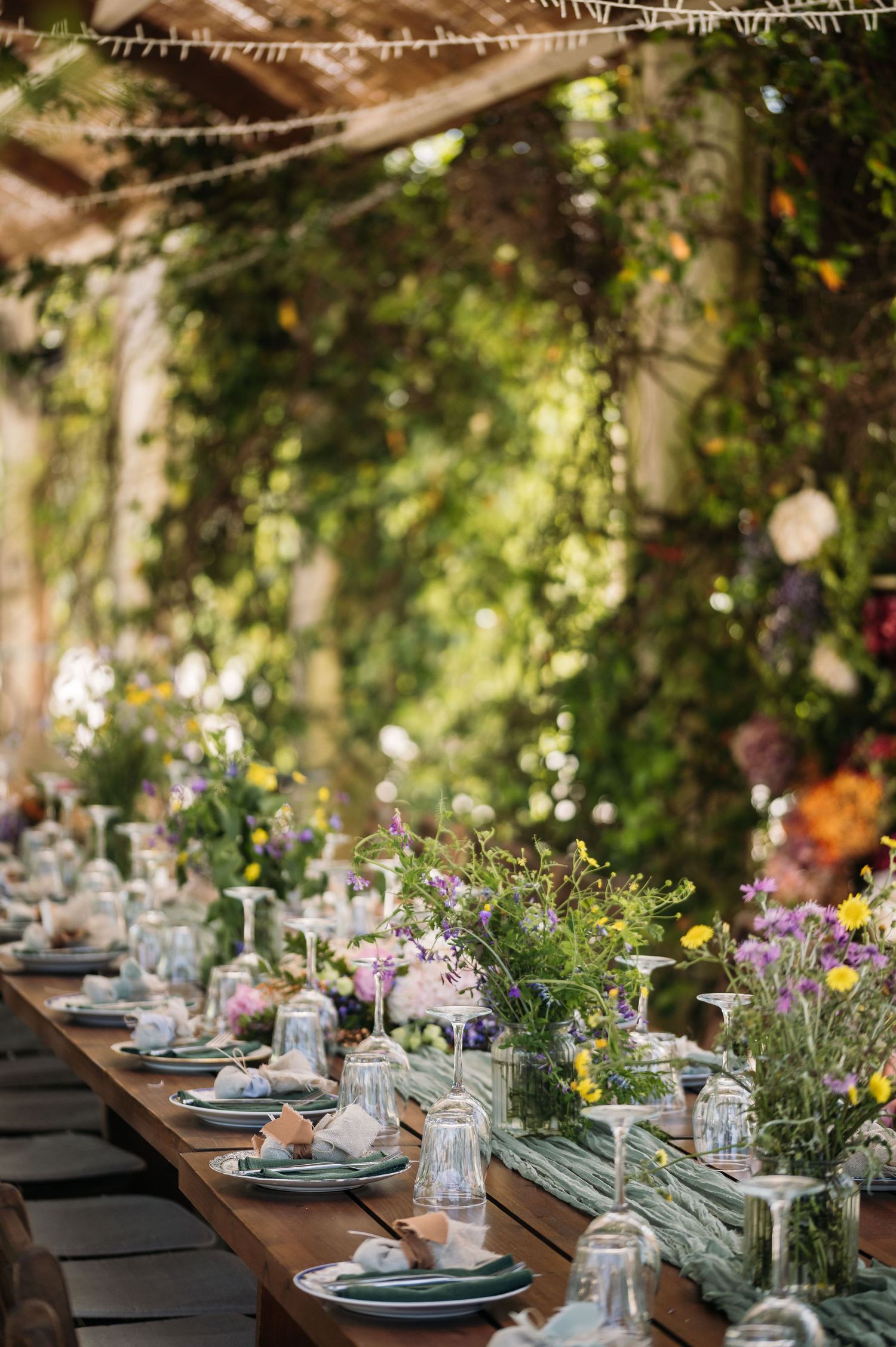 Elegant garden table setting with wildflowers and mason jars under a vine-covered pergola.
