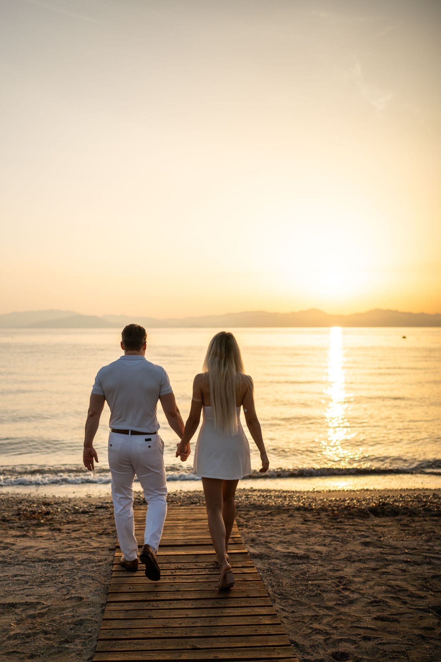 Couple walking hand in hand on a beach boardwalk during golden sunset.