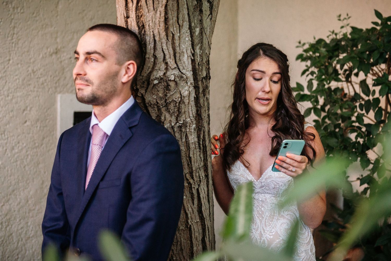 Wedding couple stands on opposite sides of tree trunk, with bride checking phone while groom looks away.