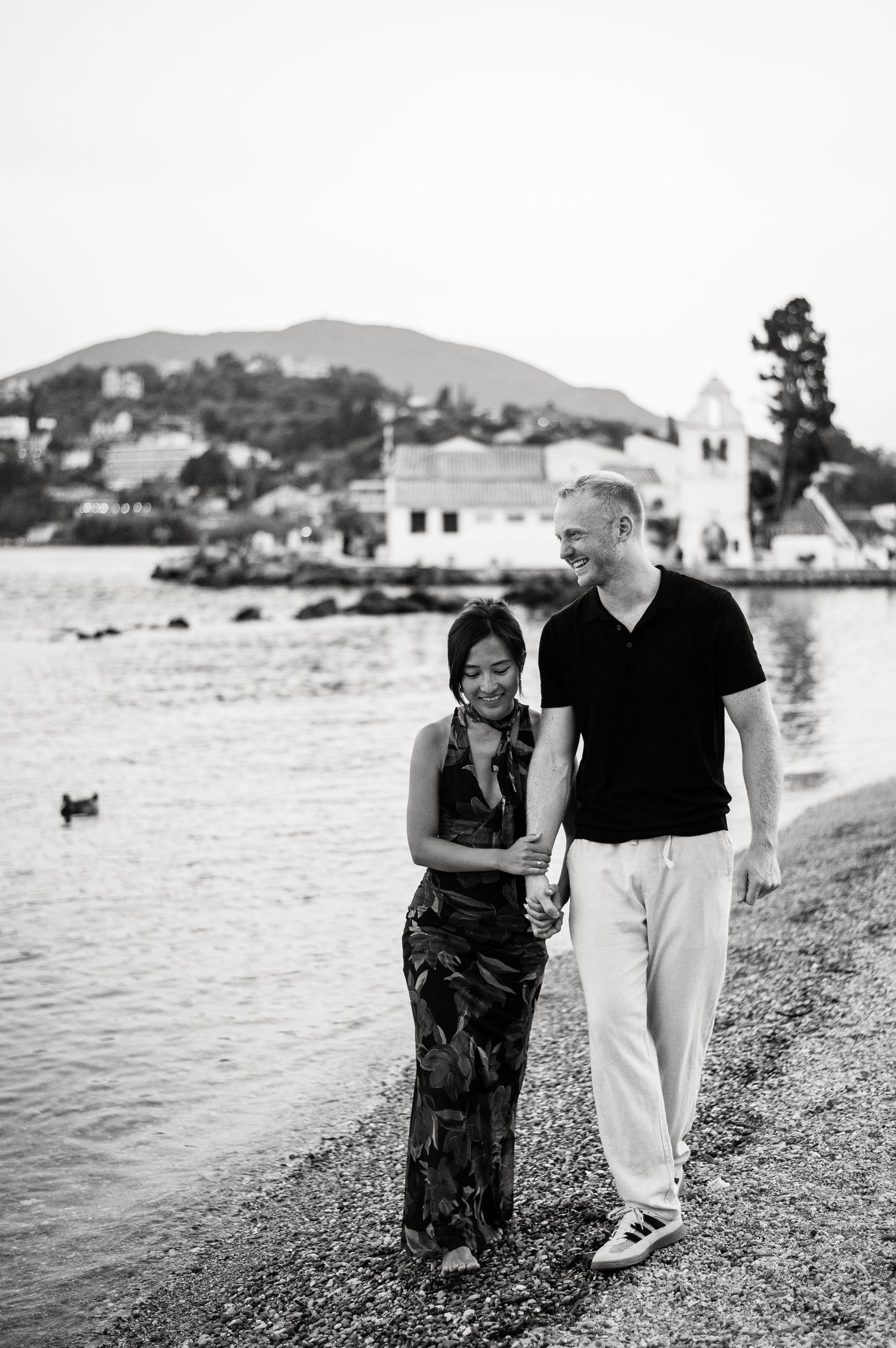 Couple walking along rocky beach with Mediterranean village in background.