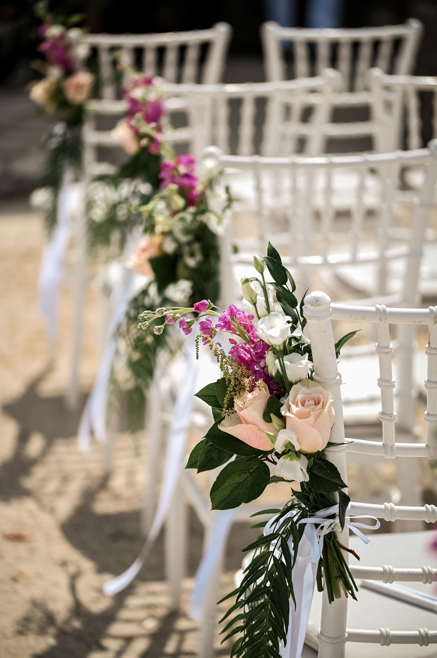 White chiavari chairs adorned with pink and white floral arrangements line a wedding ceremony aisle.