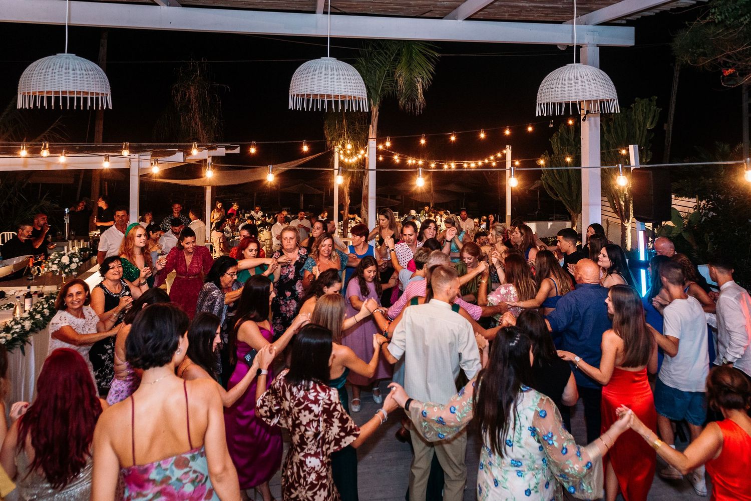 Large crowd of wedding guests dancing and celebrating on an outdoor patio under string lights.