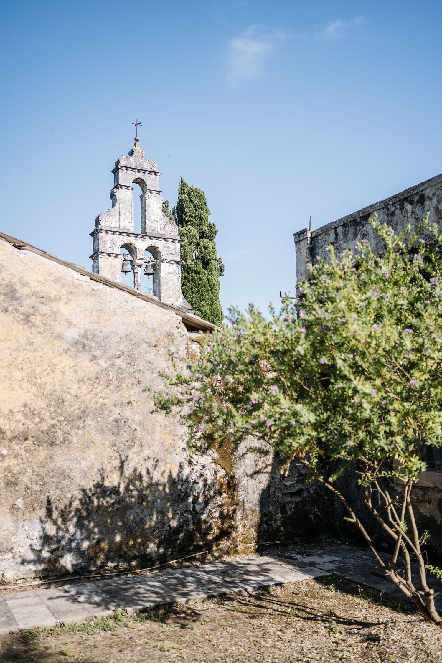 Ancient stone church bell tower rises against blue sky with green trees in Mediterranean setting.