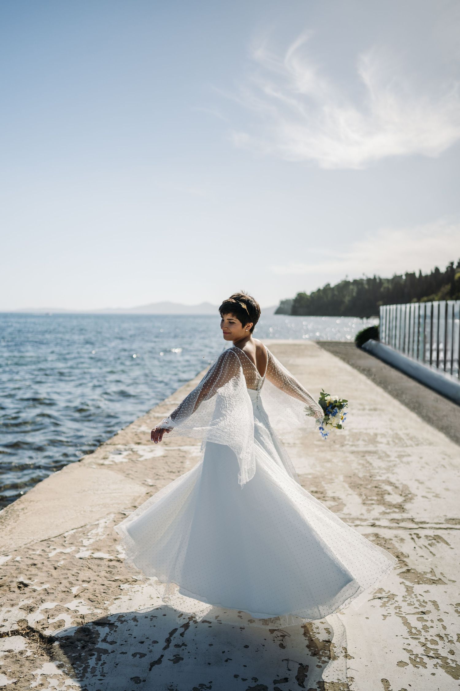 A flowing white wedding dress twirls gracefully along a seaside pier with sparkling water in the background.