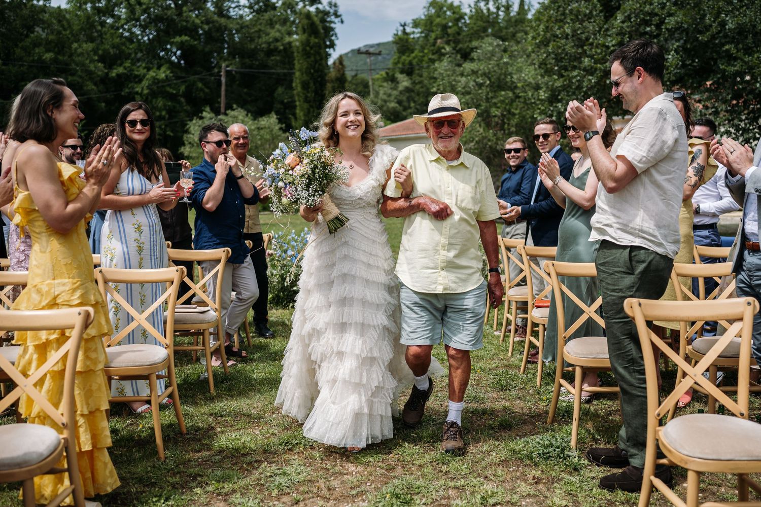 Wedding guests seated in rustic wooden chairs applaud during an outdoor garden ceremony.