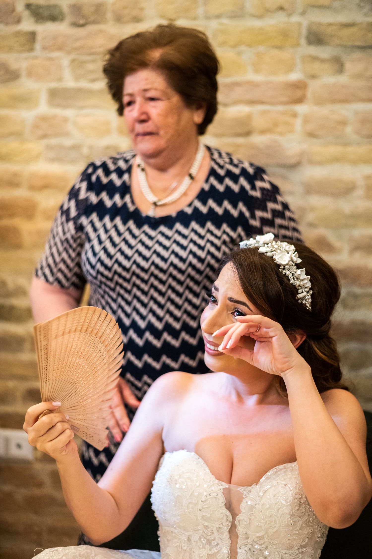 A bride in a white dress holds up a decorative wooden fan while posing against a brick wall.