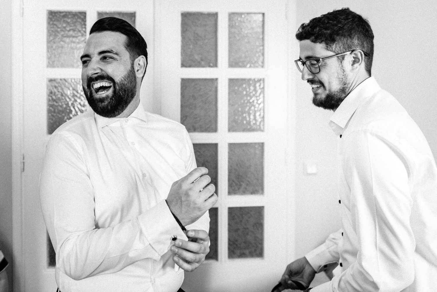 Two men in white shirts share a candid moment in front of a windowed door.