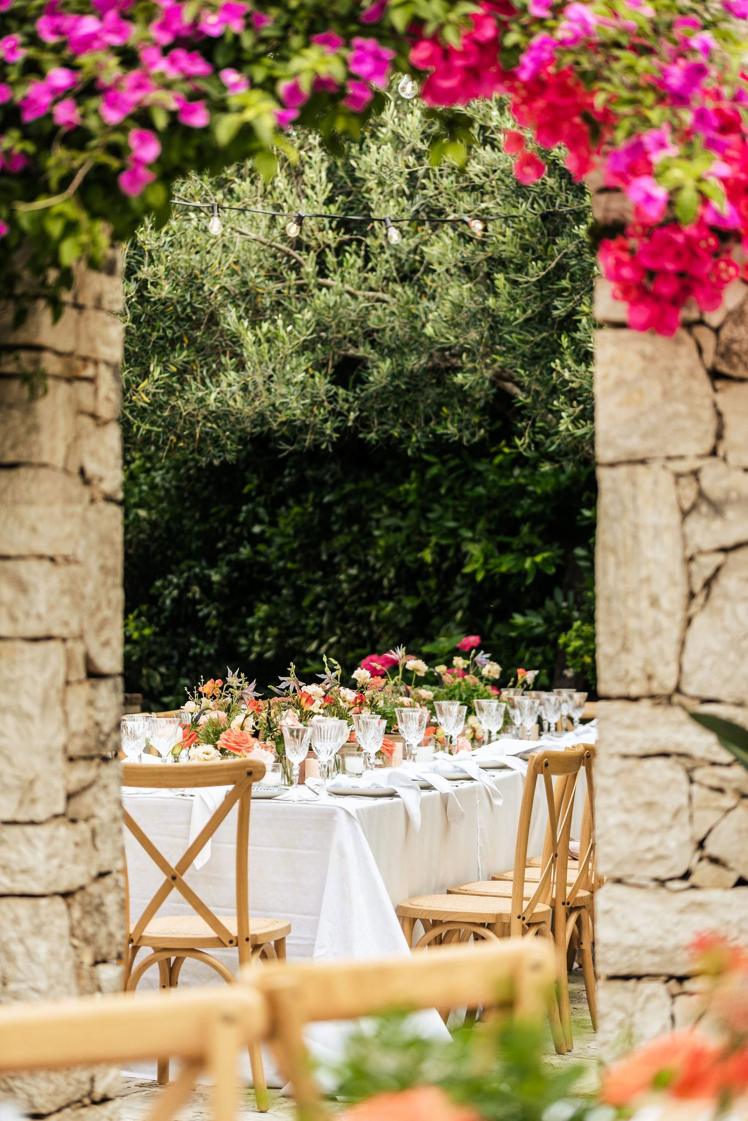 Stone archway decorated with vibrant pink flowers frames an outdoor wedding dining setup.