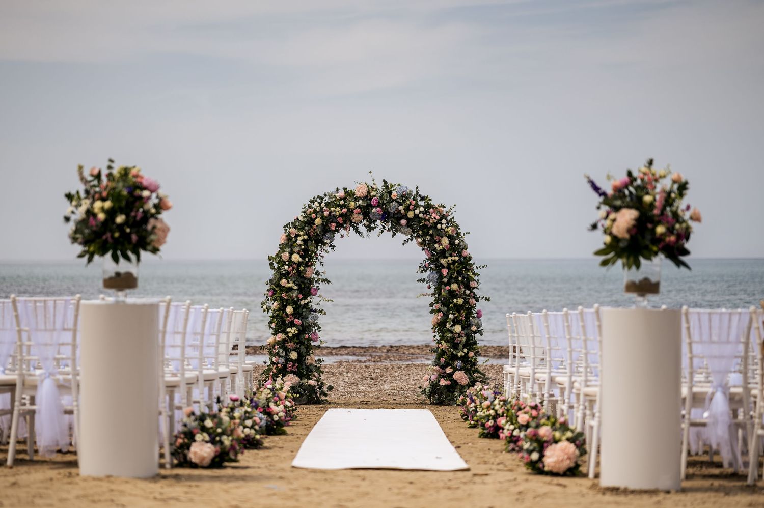 White chairs and floral arrangements line a beachside wedding ceremony setup with ocean views.