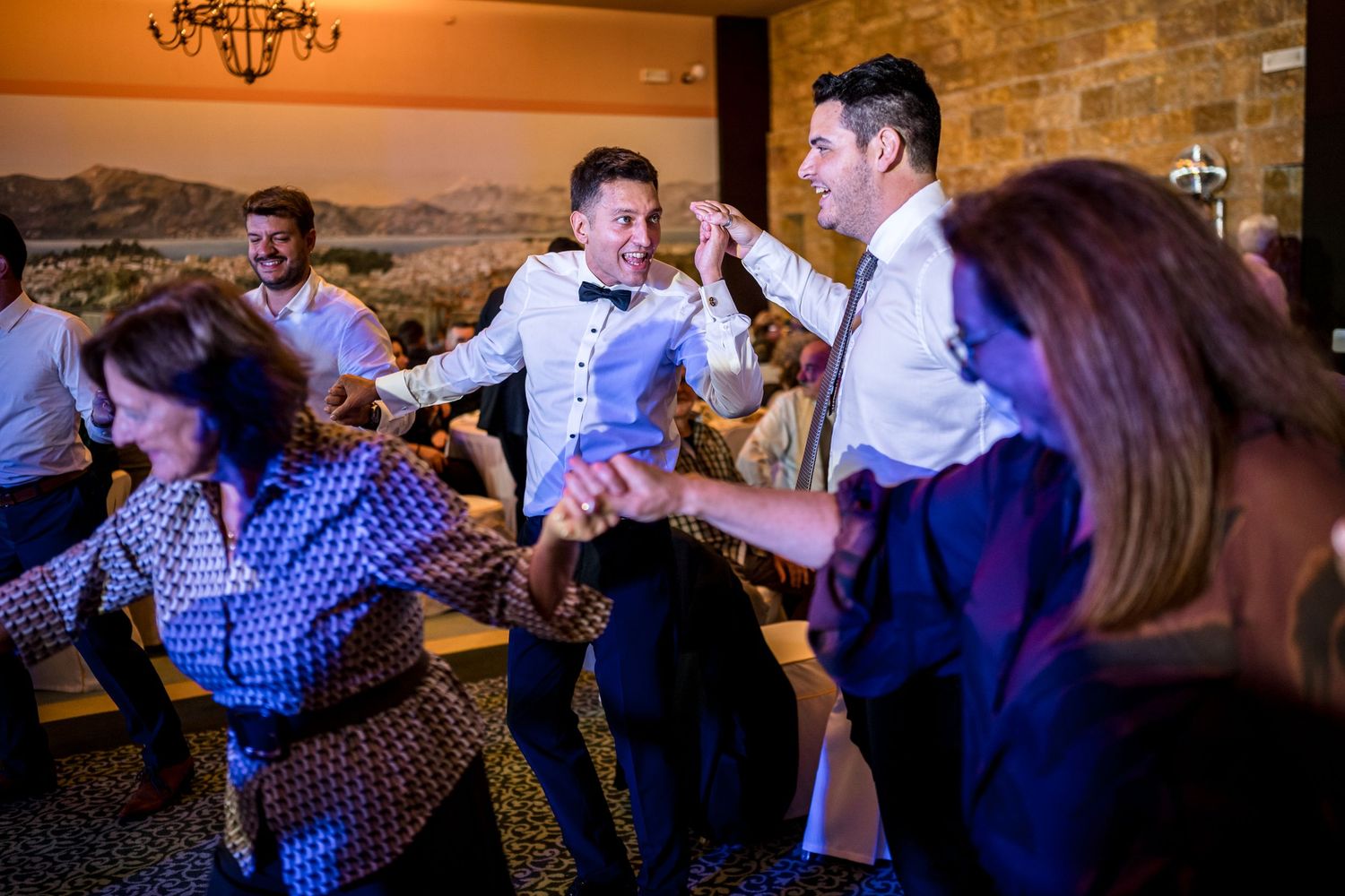 Wedding party dancing and celebrating at evening reception with warm ambient lighting.