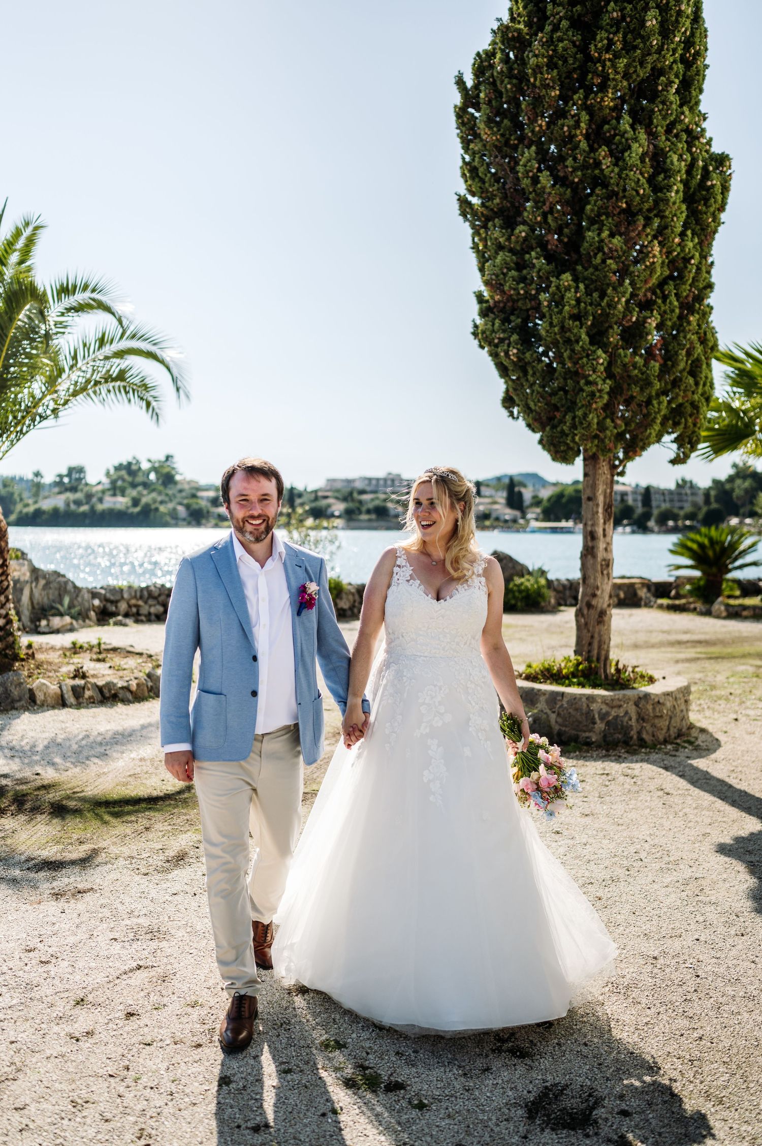 Newlyweds strolling on sunny path with cypress trees and ocean view.