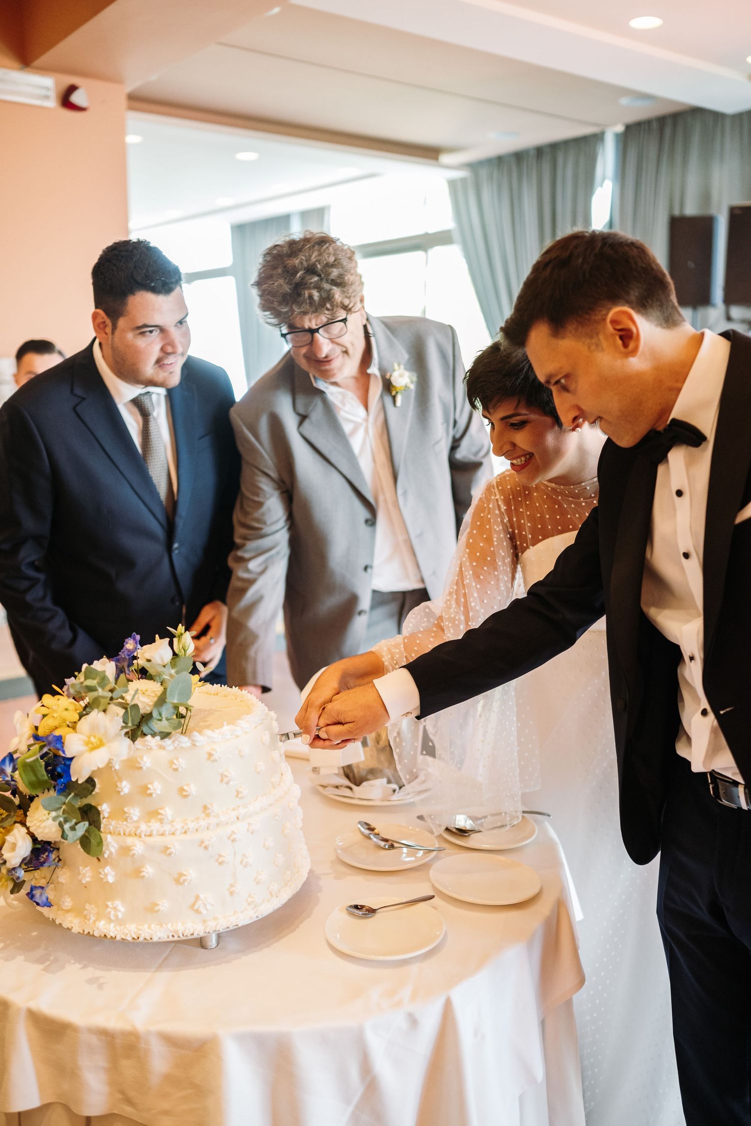 Wedding guests gather around a white tiered cake during the reception celebration.