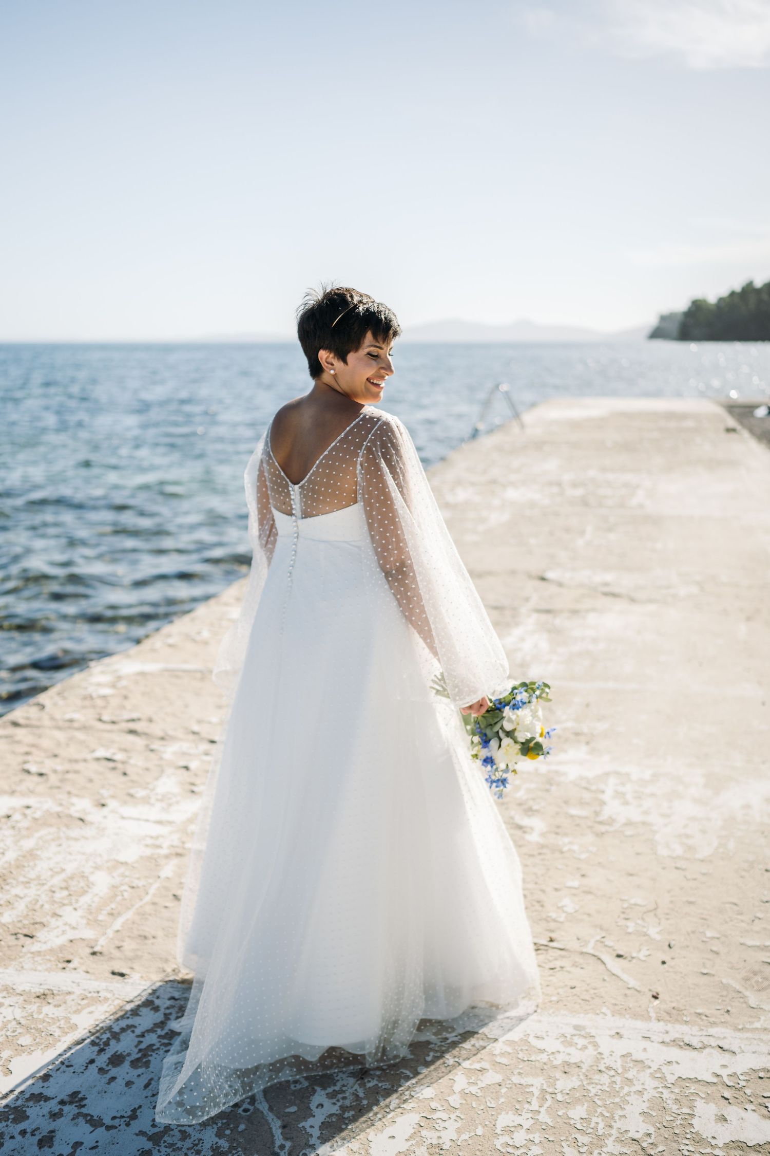 A flowing white wedding gown with sheer sleeves catches the sunlight on a coastal walkway.