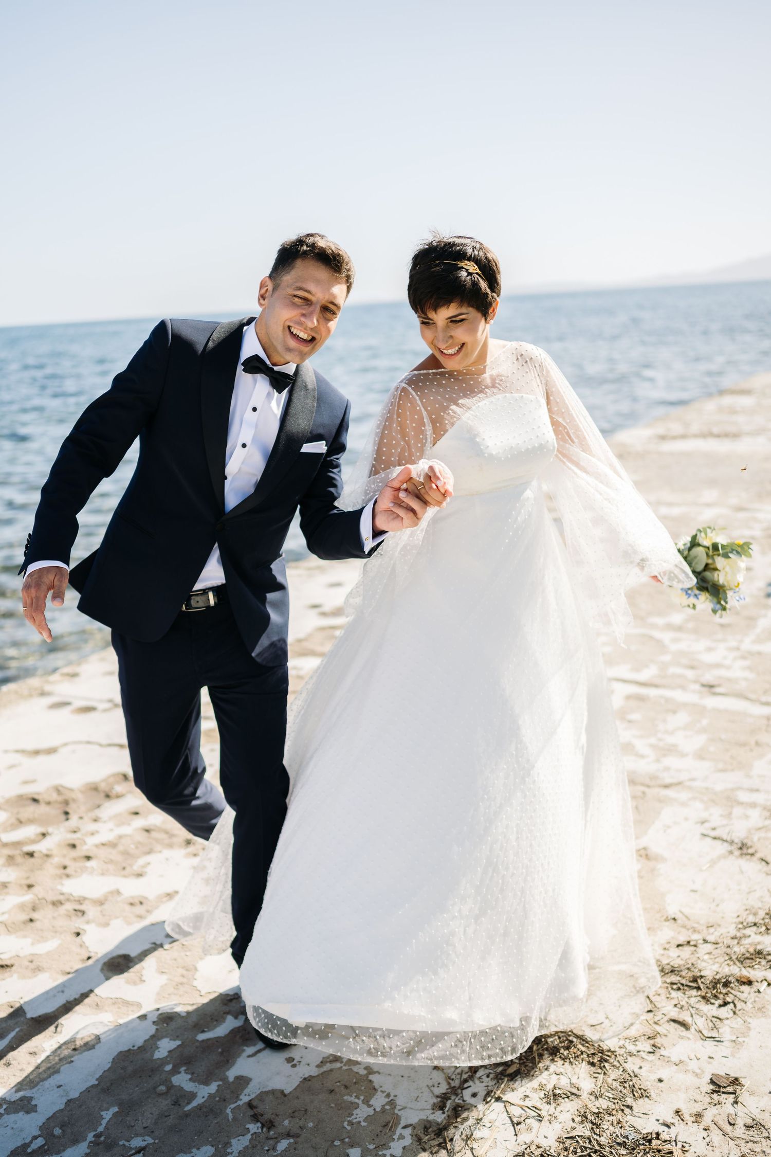 A couple in wedding attire shares lighthearted moments on a sunny beach.