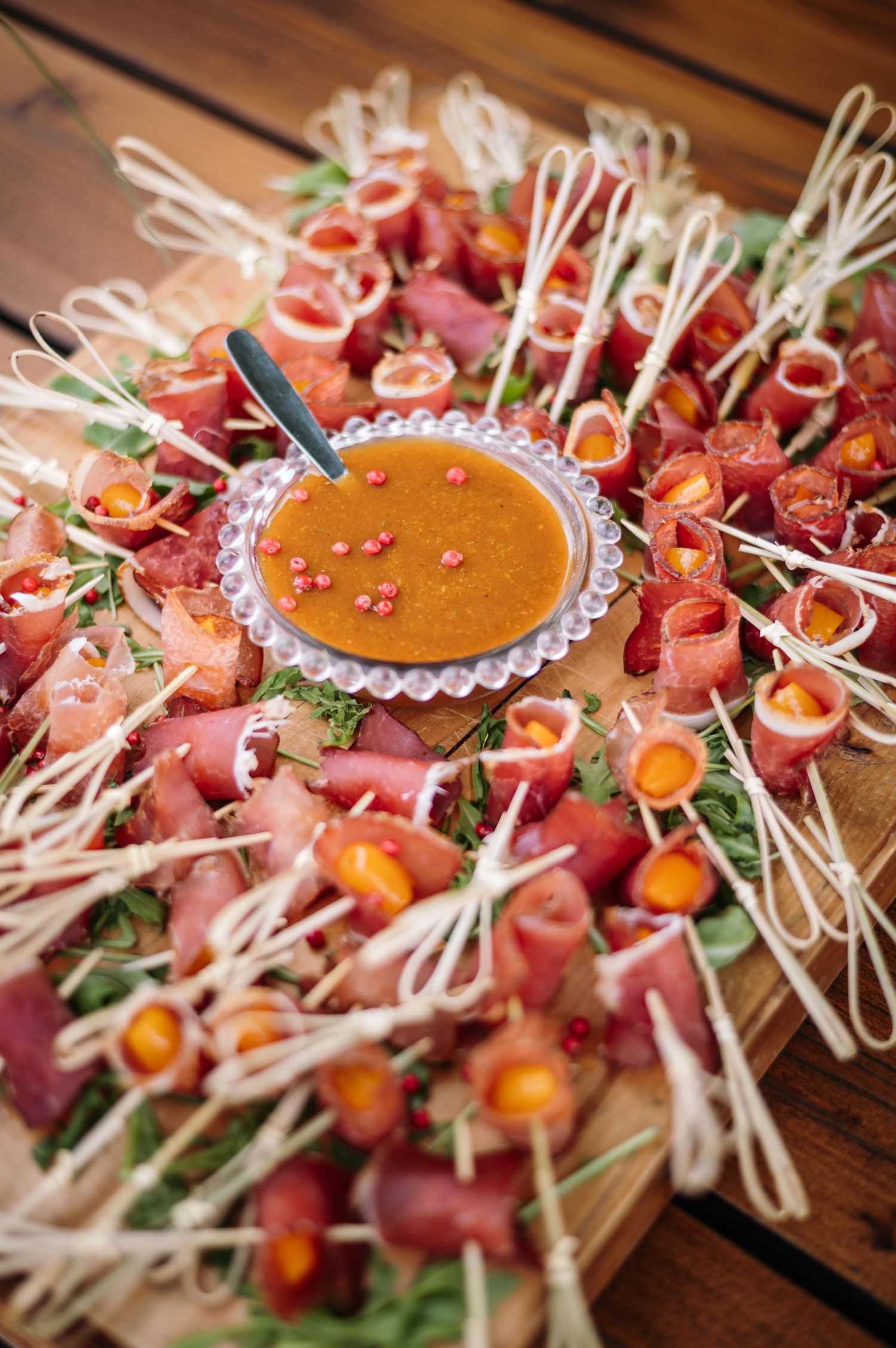 Appetizer platter with cured meats, garnishes and dipping sauce arranged on wooden surface.