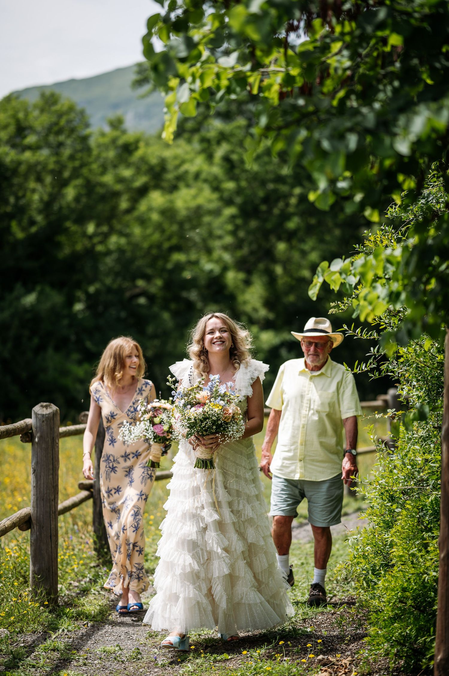 Group walking along a tree-lined path in formal attire during an outdoor celebration.