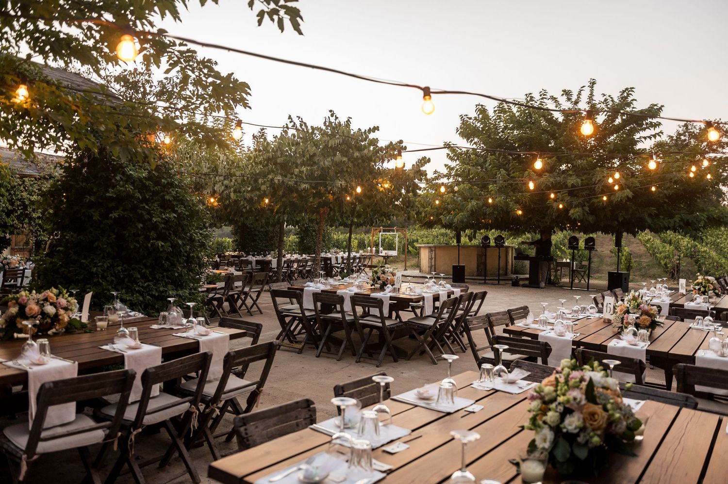 Outdoor wedding reception tables under string lights at dusk with a natural garden backdrop.