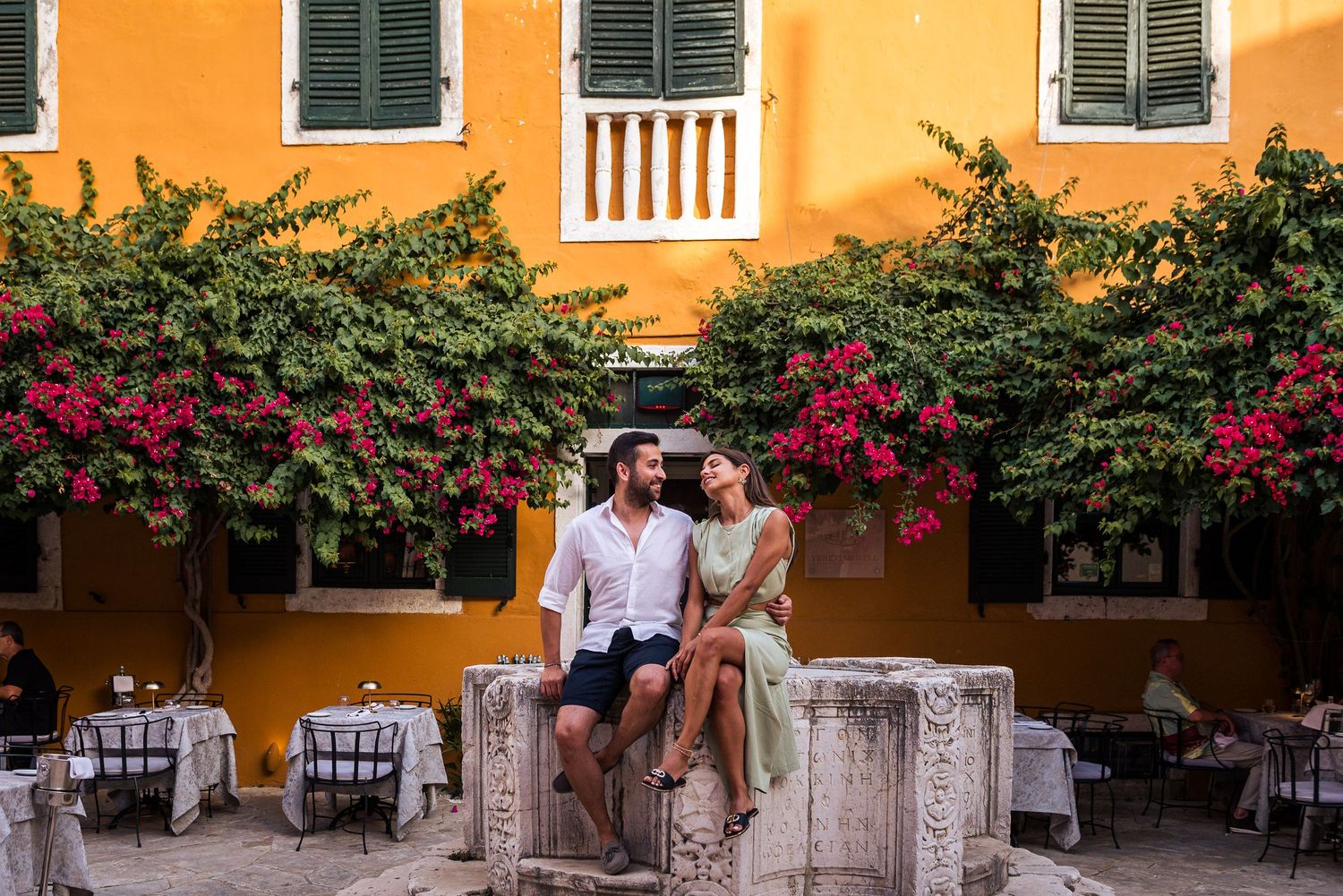 Vibrant orange Mediterranean building facade with green shutters and outdoor dining area.