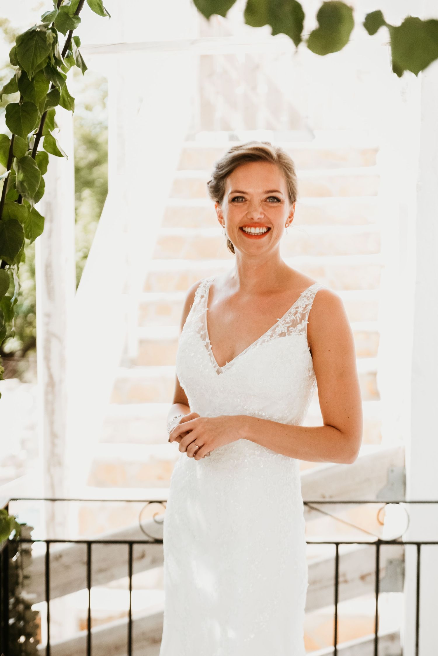 A person in a white wedding dress smiles joyfully on a balcony with green foliage.