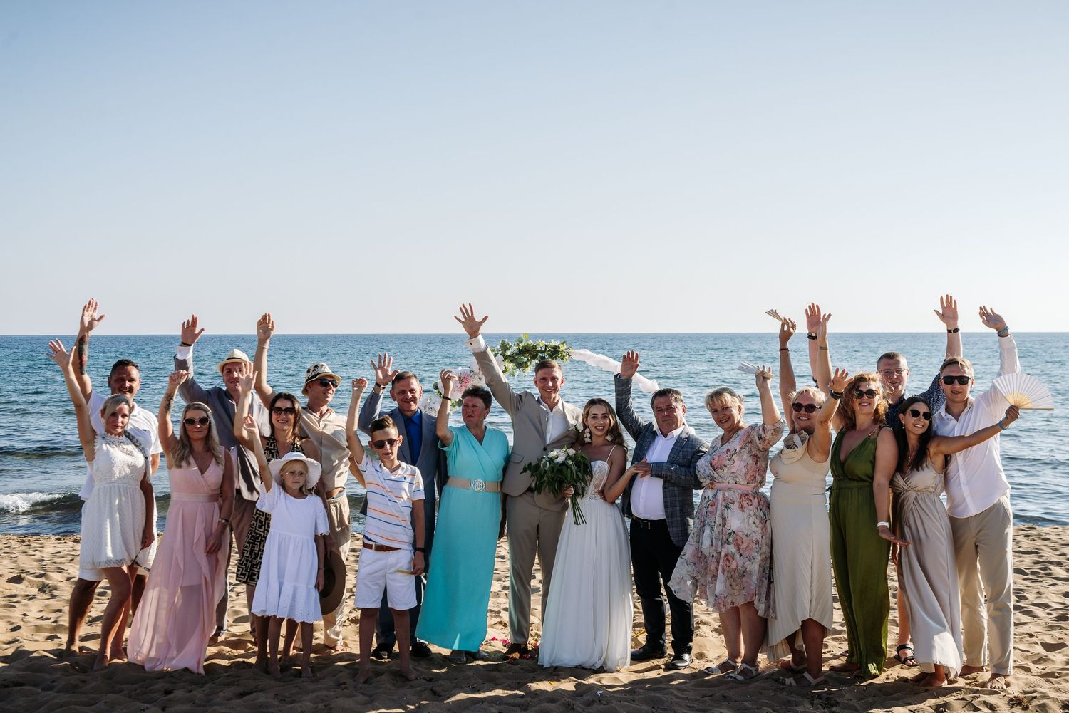 Large group photo of wedding party celebrating on beach with arms raised.