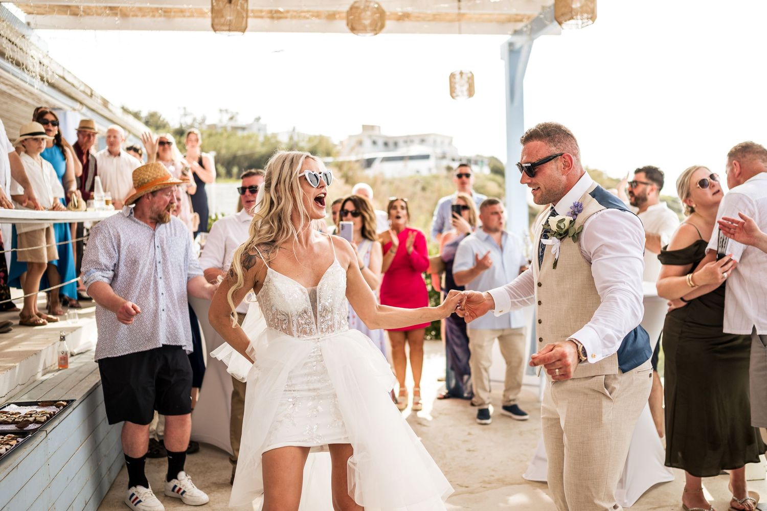 Wedding reception guests dancing and mingling under an open-air pavilion with ocean views.