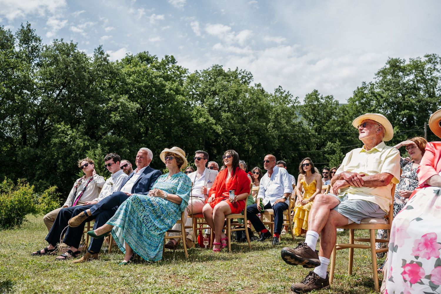 Wedding guests enjoy a sunny outdoor ceremony with wooden chairs on a grassy hillside.