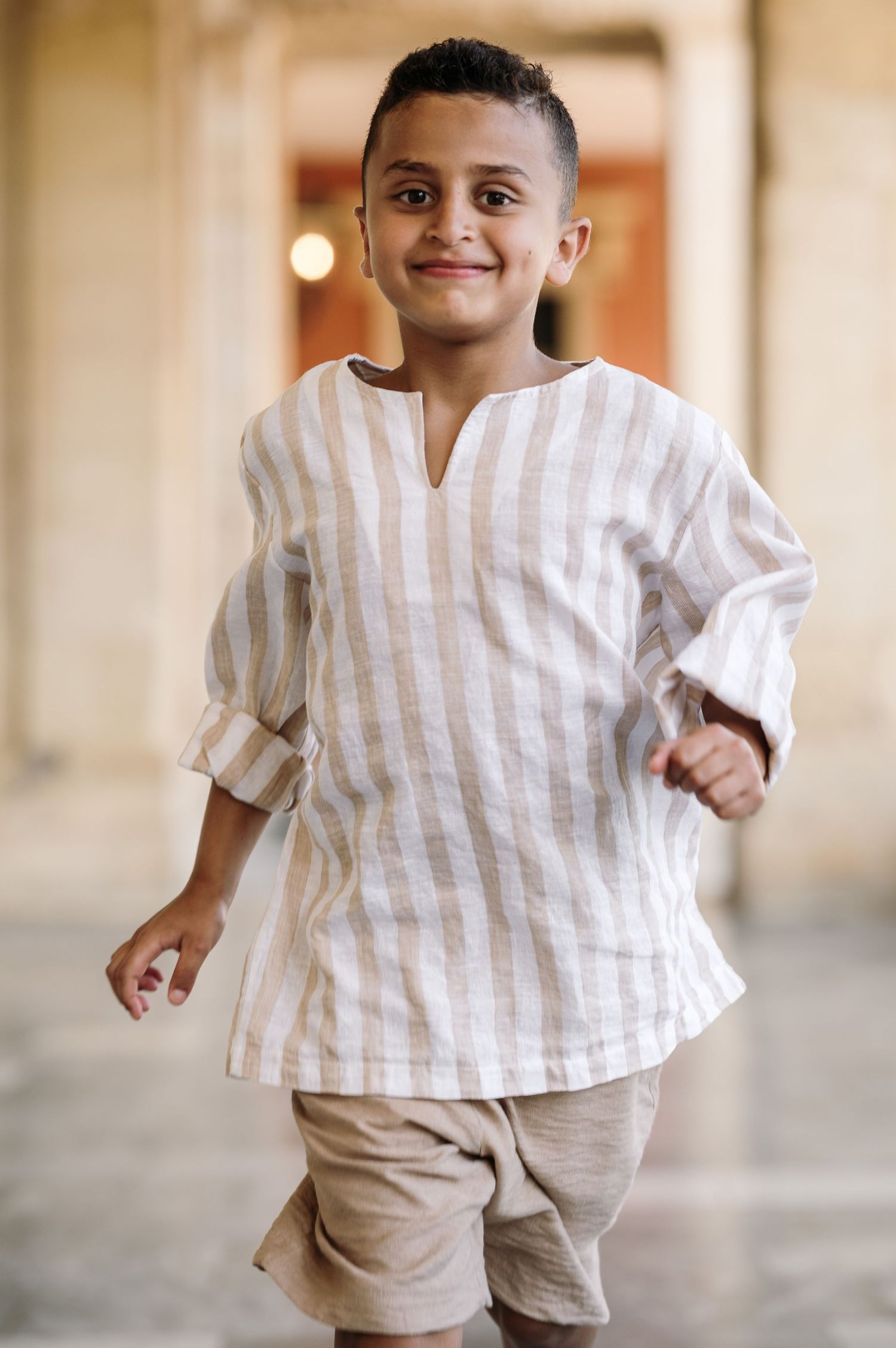 Child wearing striped linen outfit runs playfully through a sunlit corridor.
