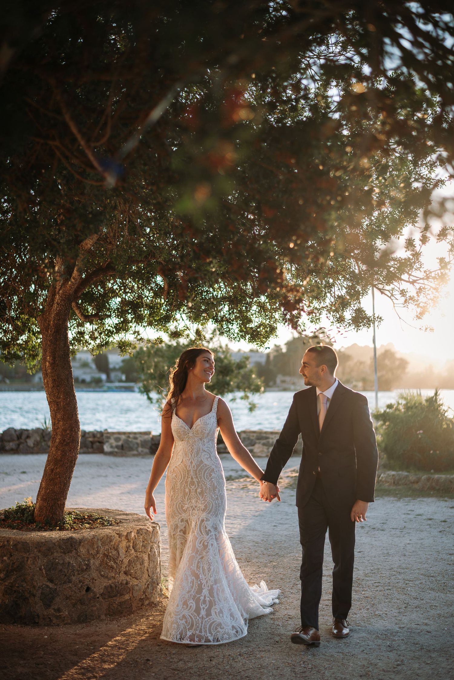 A couple in wedding attire walks hand in hand under a tree during golden hour at sunset.