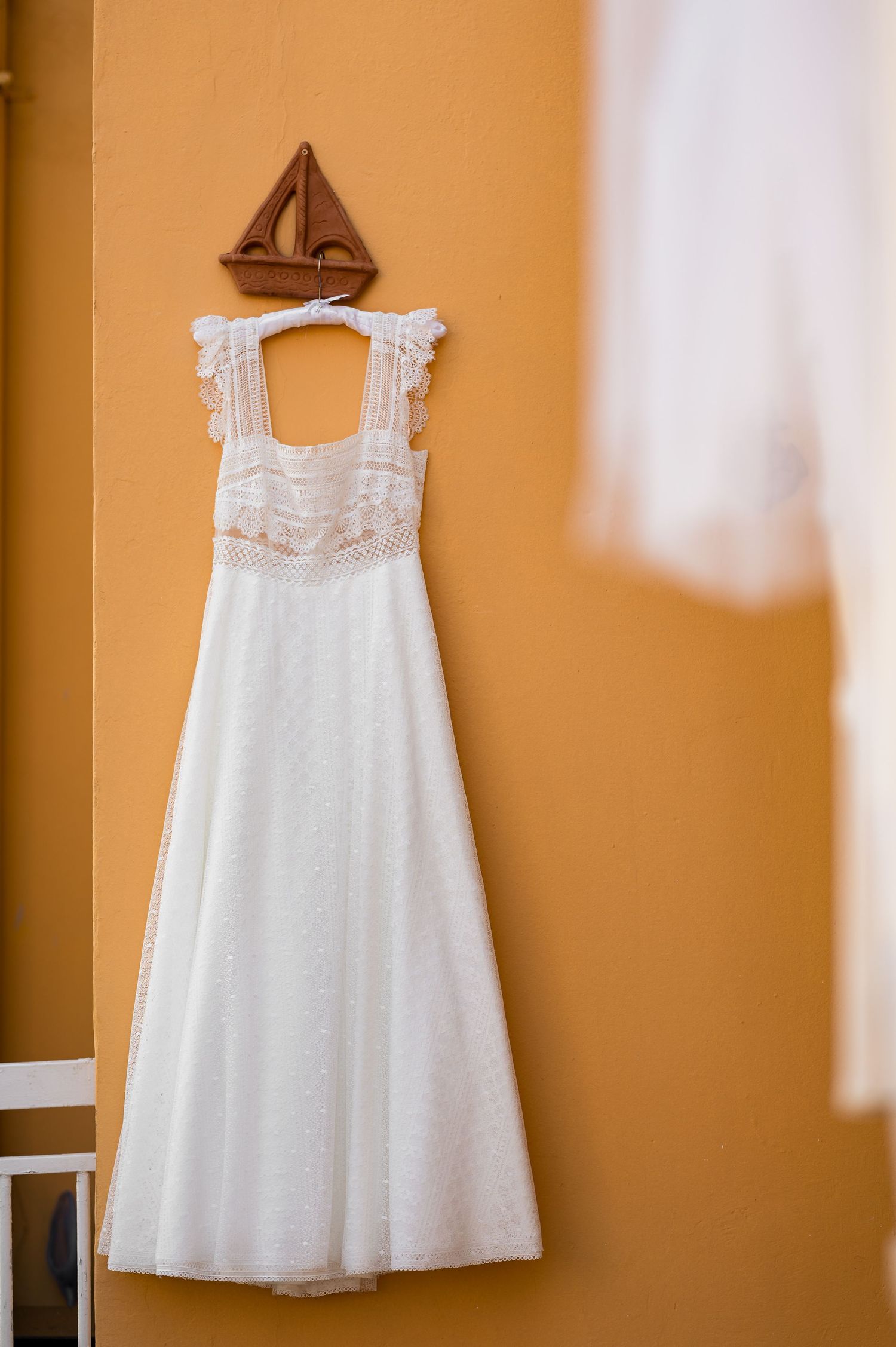 A white wedding dress with lace details hangs on a wall with a wooden sailboat decoration above it.