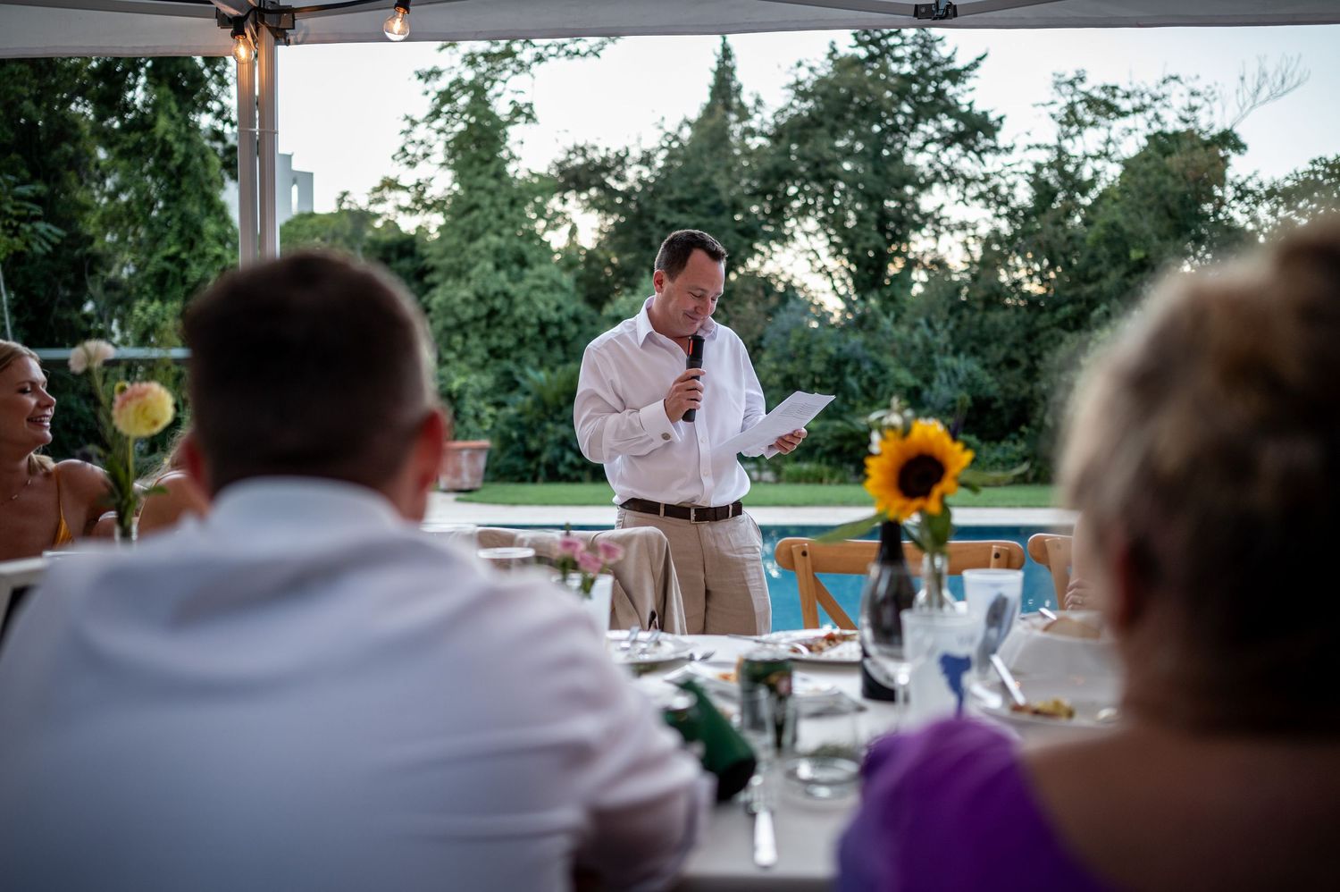 Wedding reception guests seated at tables under white tent with sunflowers visible.
