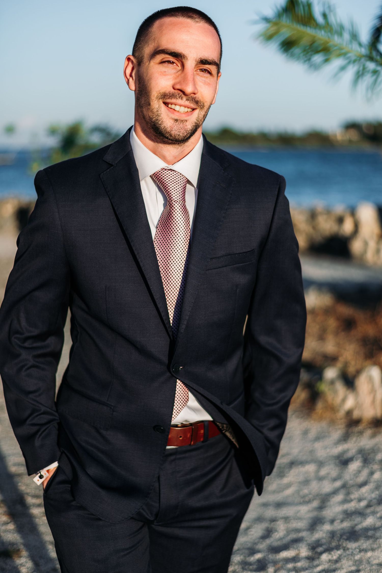 Groom in classic black suit and pink tie poses on tropical beach.