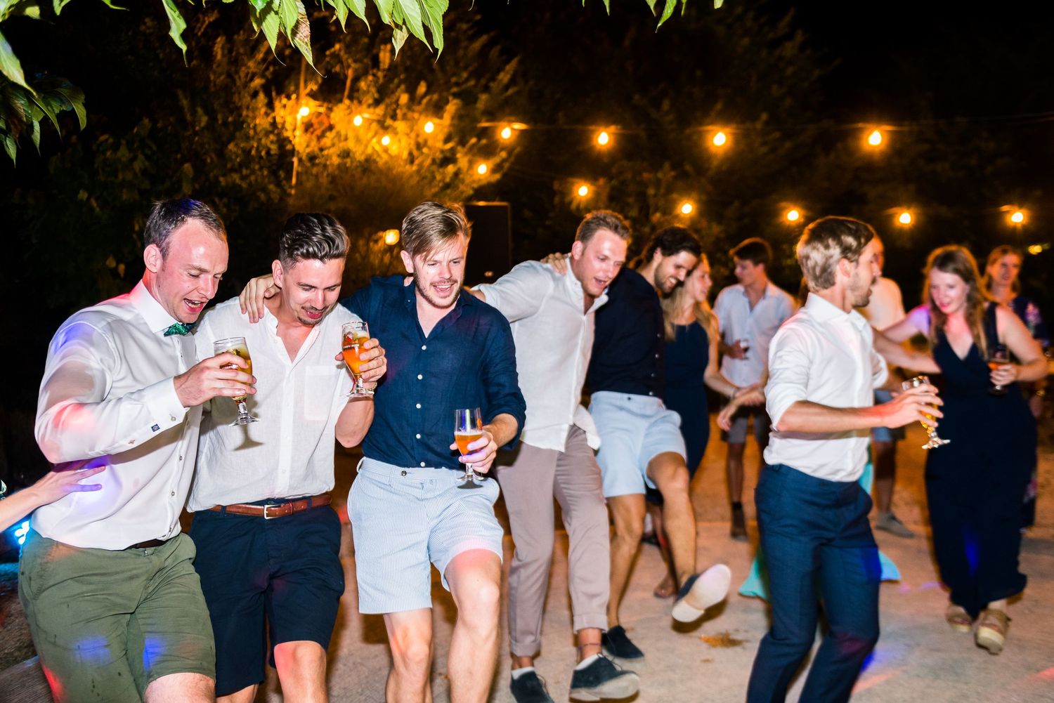 Wedding guests dance in casual attire under warm outdoor lighting at night.
