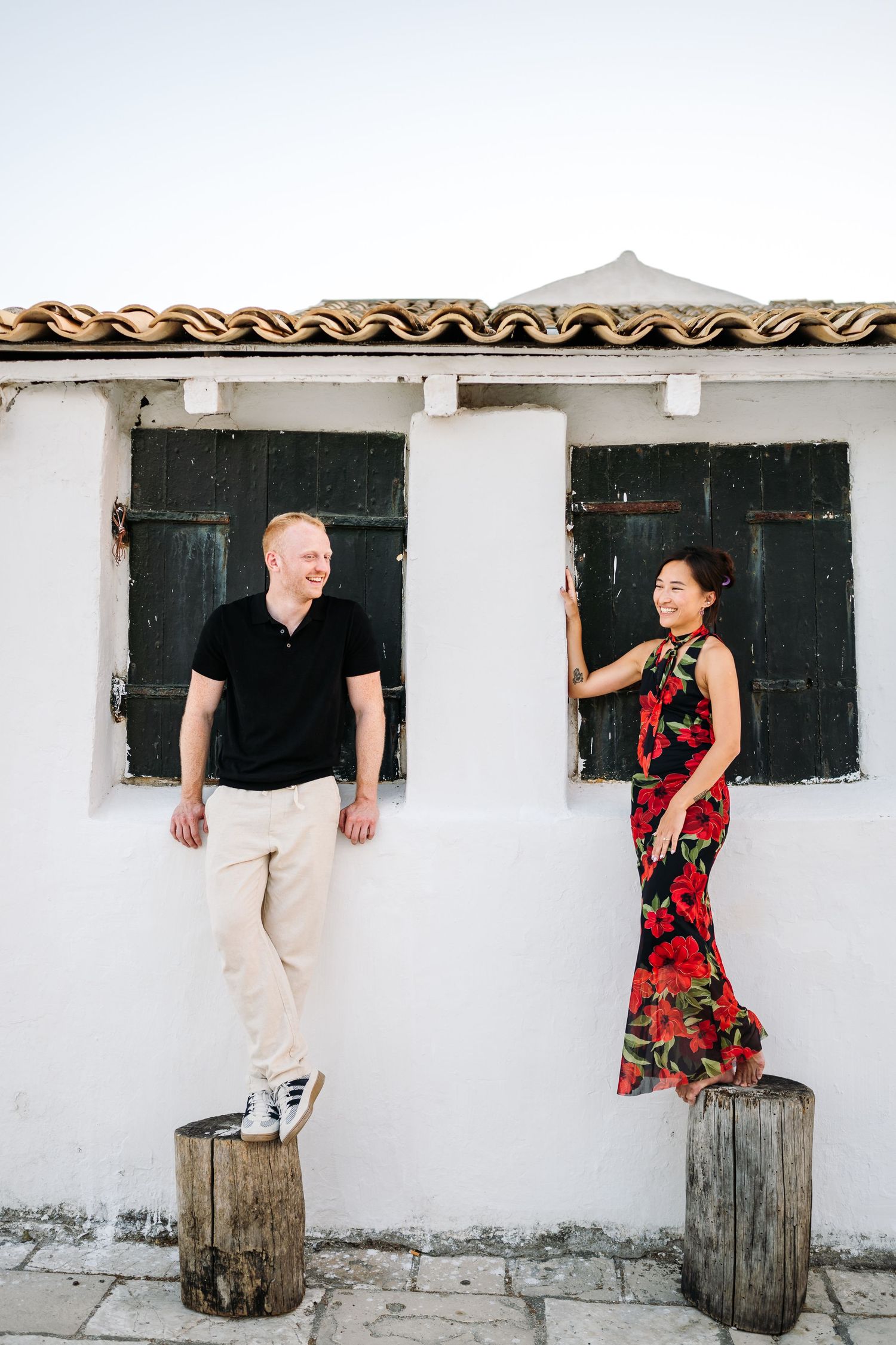 Couple posing playfully on wooden barrels against white wall with dark windows.