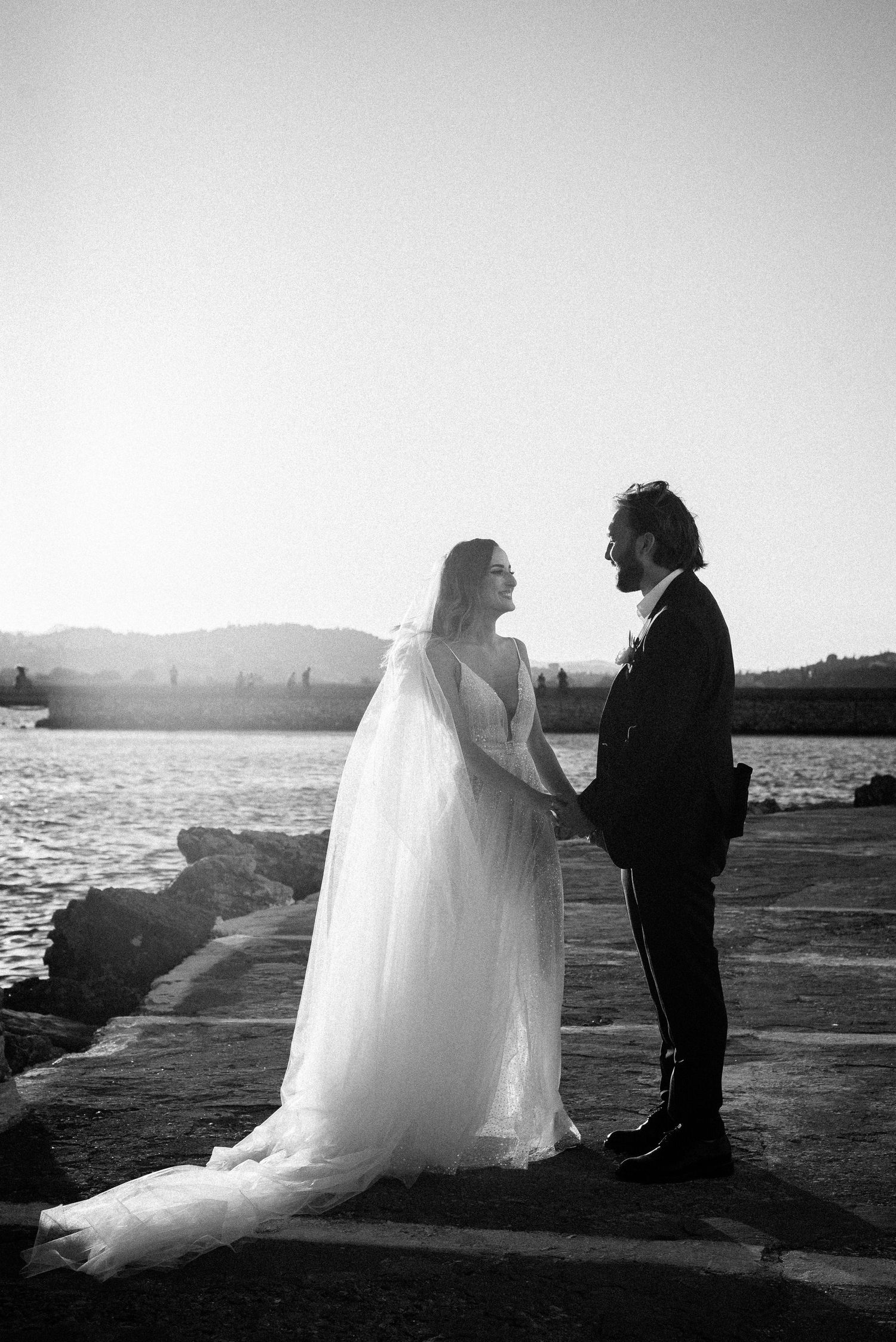 Silhouetted wedding couple poses against dramatic sunset by the water.