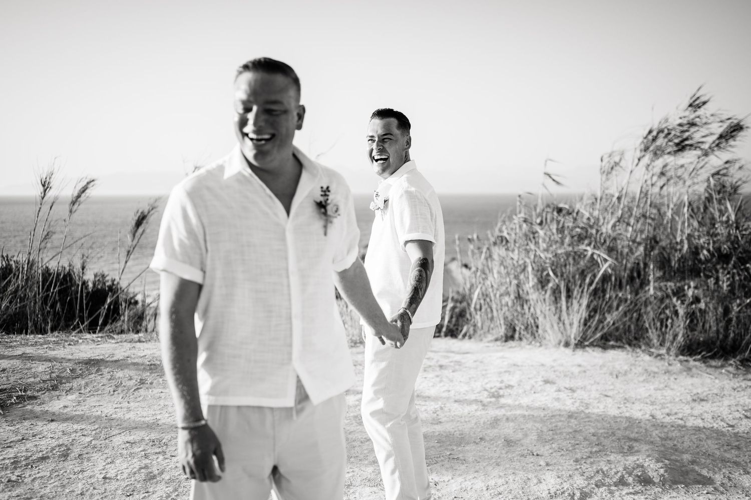 Black and white portrait of couple in white outfits standing on coastal path.
