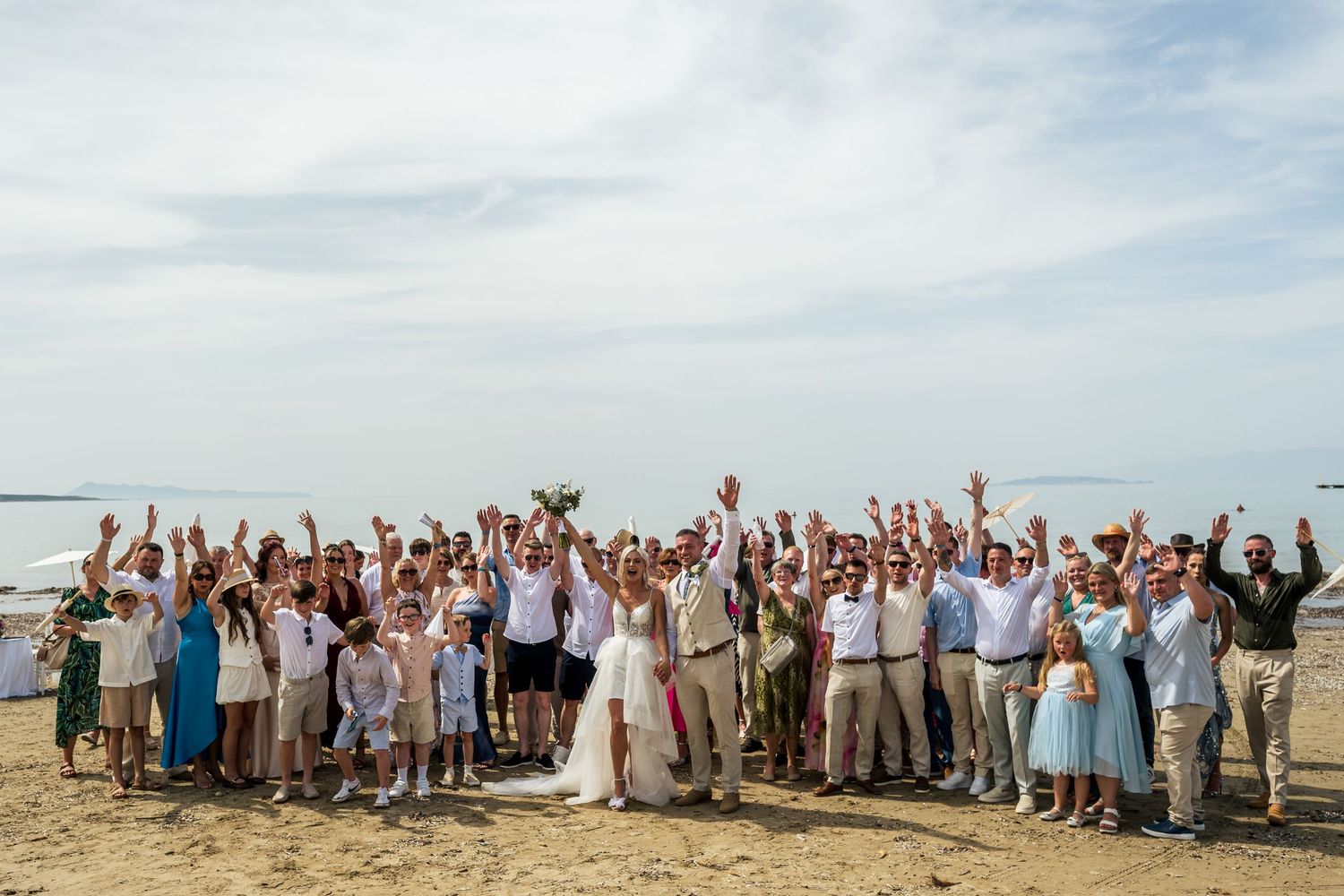 Large wedding party celebrating on a beach with arms raised in celebration.